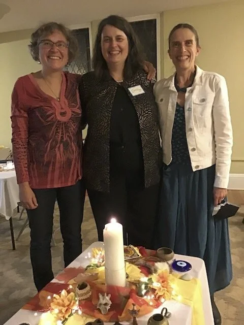 Three women standing together at a celebration, smiling, with a table decorated with a large candle, flowers, and party decorations in front of them.