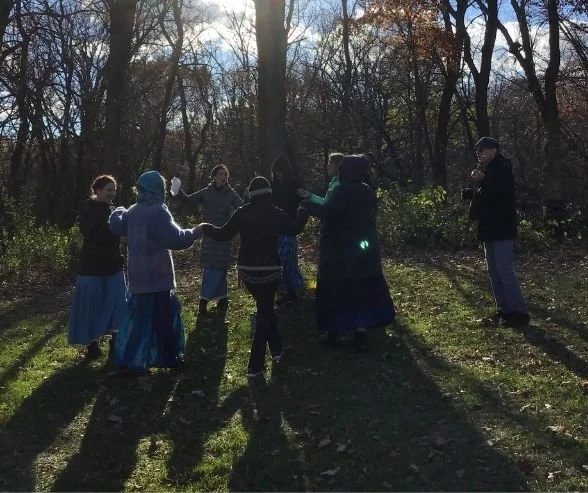 A group of children and an adult holding hands in a circle outdoors in a wooded area during daylight.
