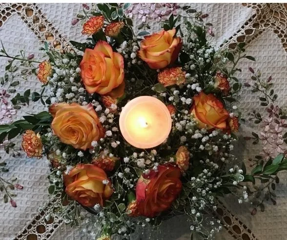 A floral arrangement with orange and yellow roses, baby's breath, and greenery surrounding a lit candle on a lace tablecloth.