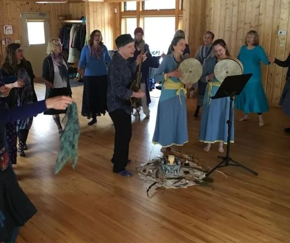 Group of people performing traditional music and dance in a wooden-paneled room, with some holding percussion instruments and others dancing.