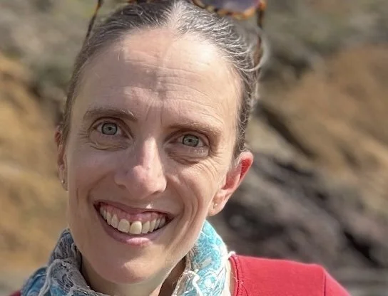 Close-up of a smiling woman with light-colored hair tied back, wearing sunglasses on her head and a red jacket outdoors.