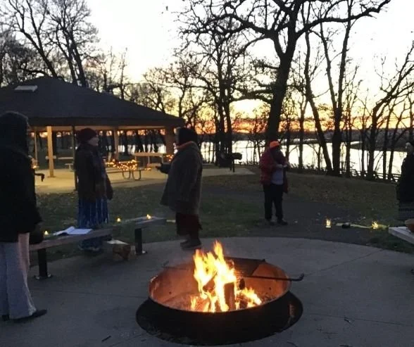 People gathered around a fire pit near a lake at sunset, with trees and a pavilion in the background.