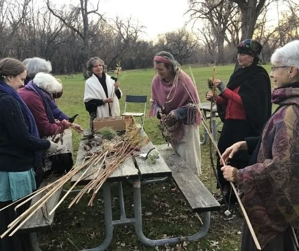 Group of women outdoors preparing and handling woven natural fiber bundles, possibly for a craft or ritual, around a picnic table with sticks and plant materials, in a park during autumn.