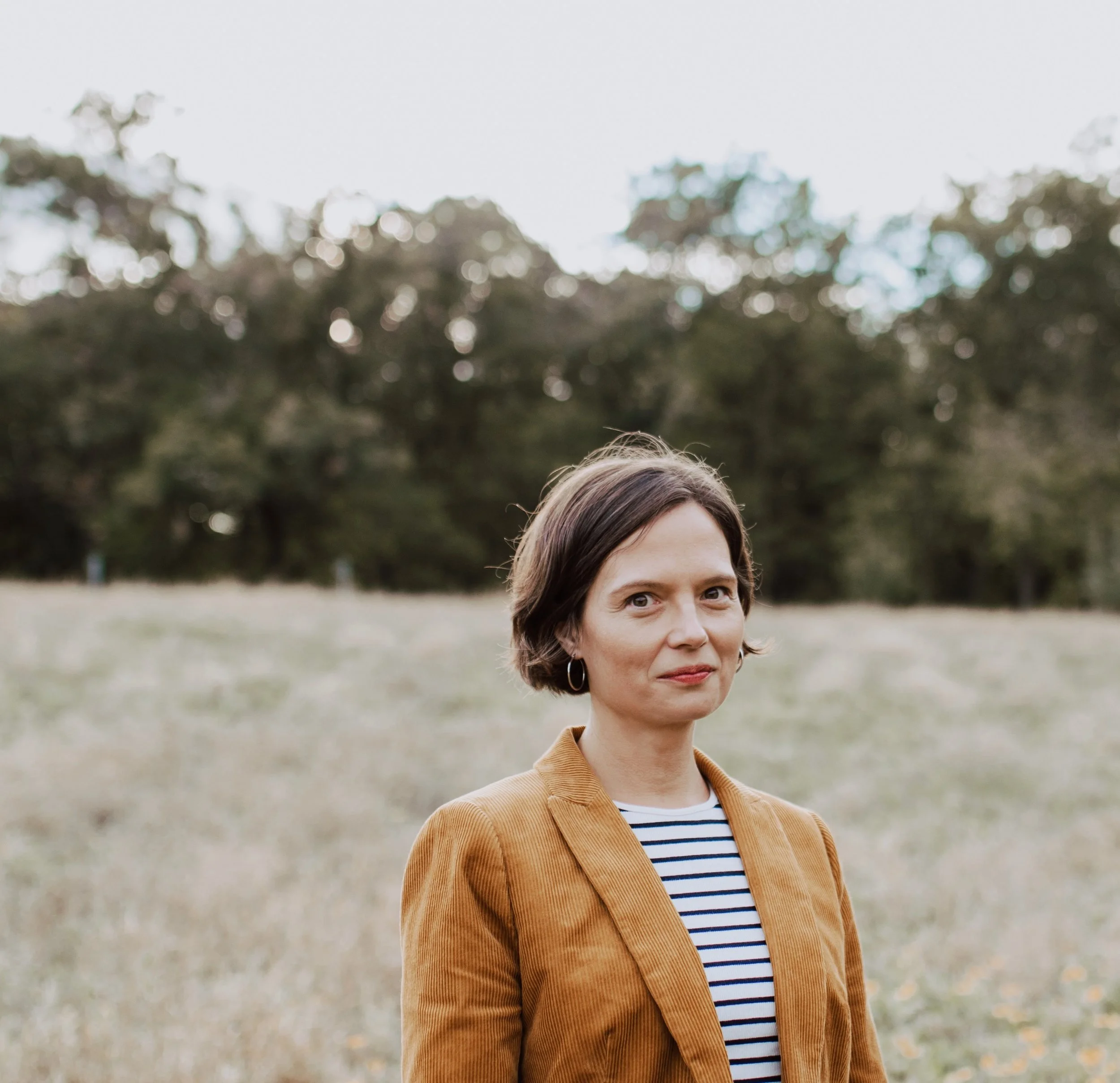 A woman with short brown hair wearing a tan blazer and striped shirt standing outdoors in a grassy field with trees in the background.