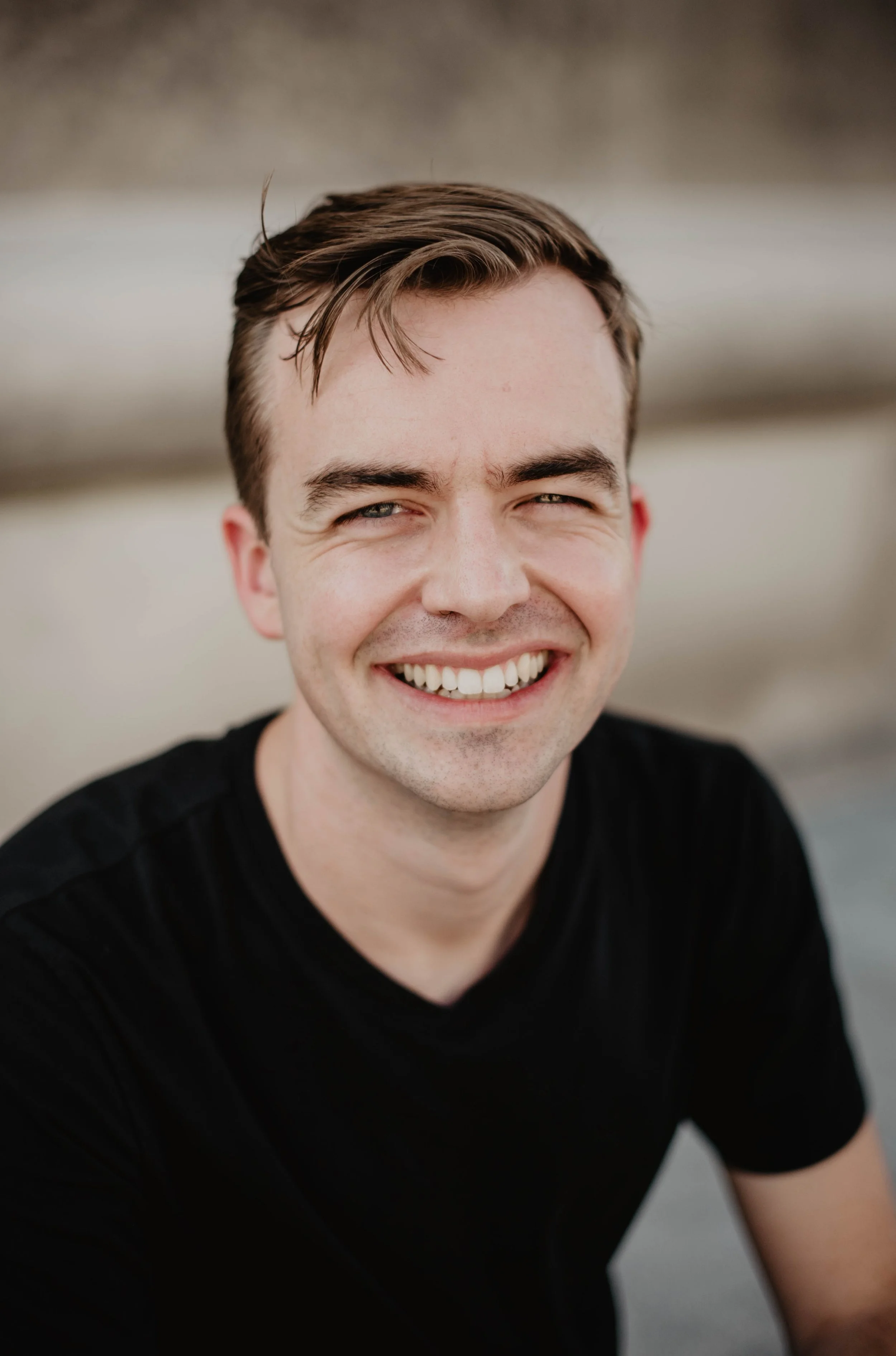 Close-up of smiling young man with short brown hair, black shirt, outdoors with blurred background.