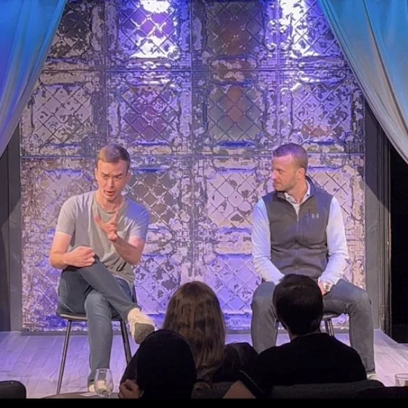 Two men are seated on a stage with a decorative wall behind them, engaged in a conversation, while an audience sits watching them.