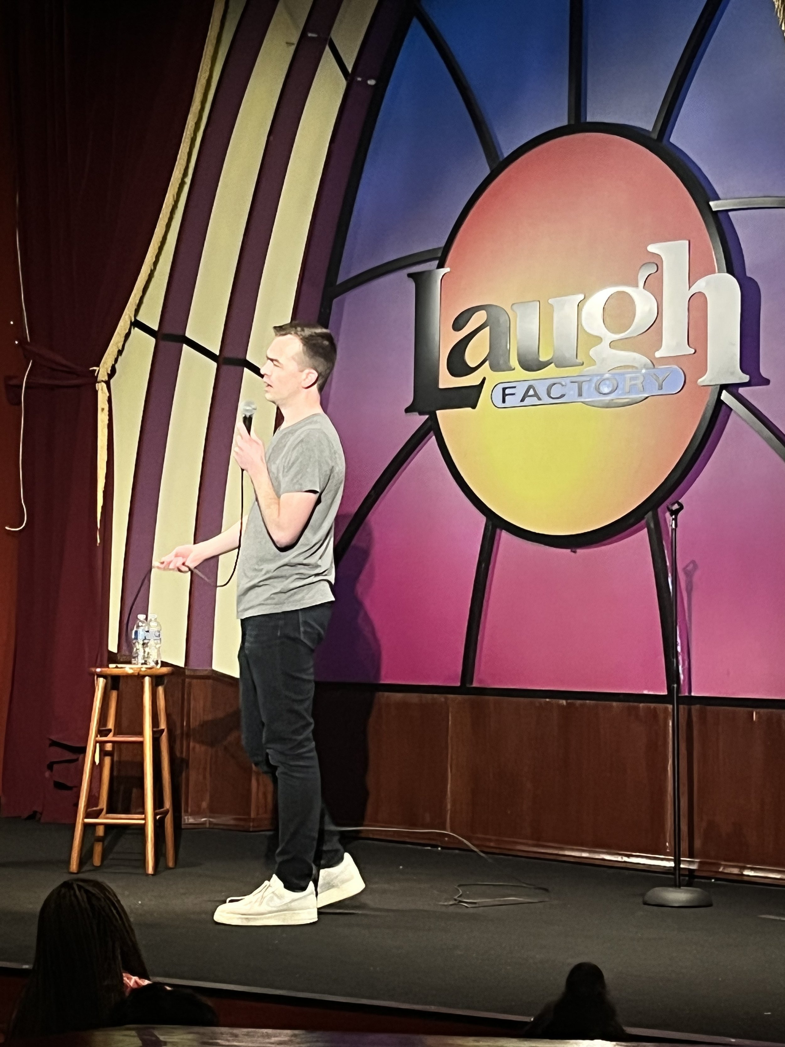 A man on stage at the Laugh Factory comedy club, holding a microphone, wearing a gray t-shirt and white sneakers. Behind him is a colorful sign with the Laugh Factory logo and a small table with water bottles. The stage has a dark floor with a black microphone stand.