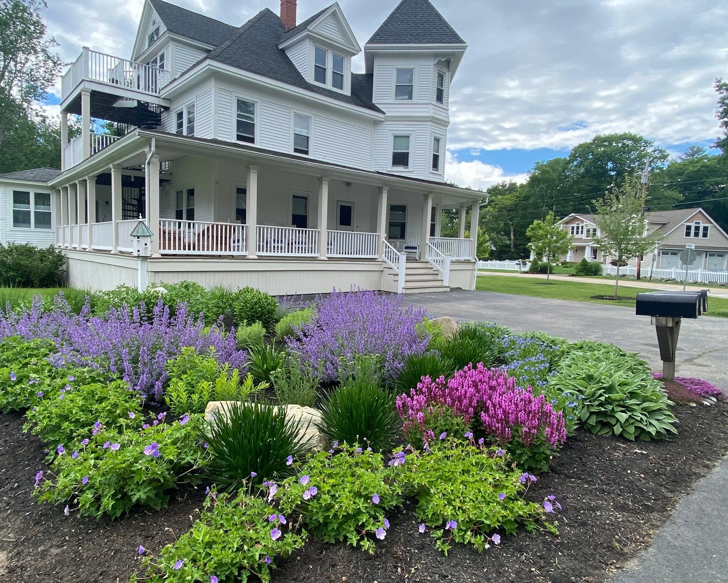 After new garden installation. A white house with a large front porch, multiple stories, and a turret, surrounded by a garden with purple, pink, and green flowering plants, a gravel driveway, and a small tree.