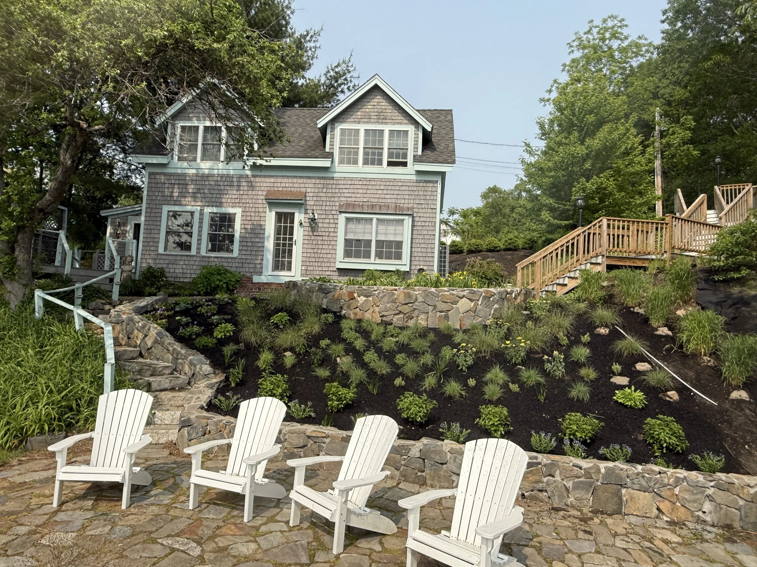 A two-story house with grey shingles, white trim, and multiple windows, situated on a hillside. The yard has a stone retaining wall, an array of green plants, and a stone patio with four white Adirondack chairs, and a wooden staircase leading to a deck on the upper level. Tall trees surround the house.