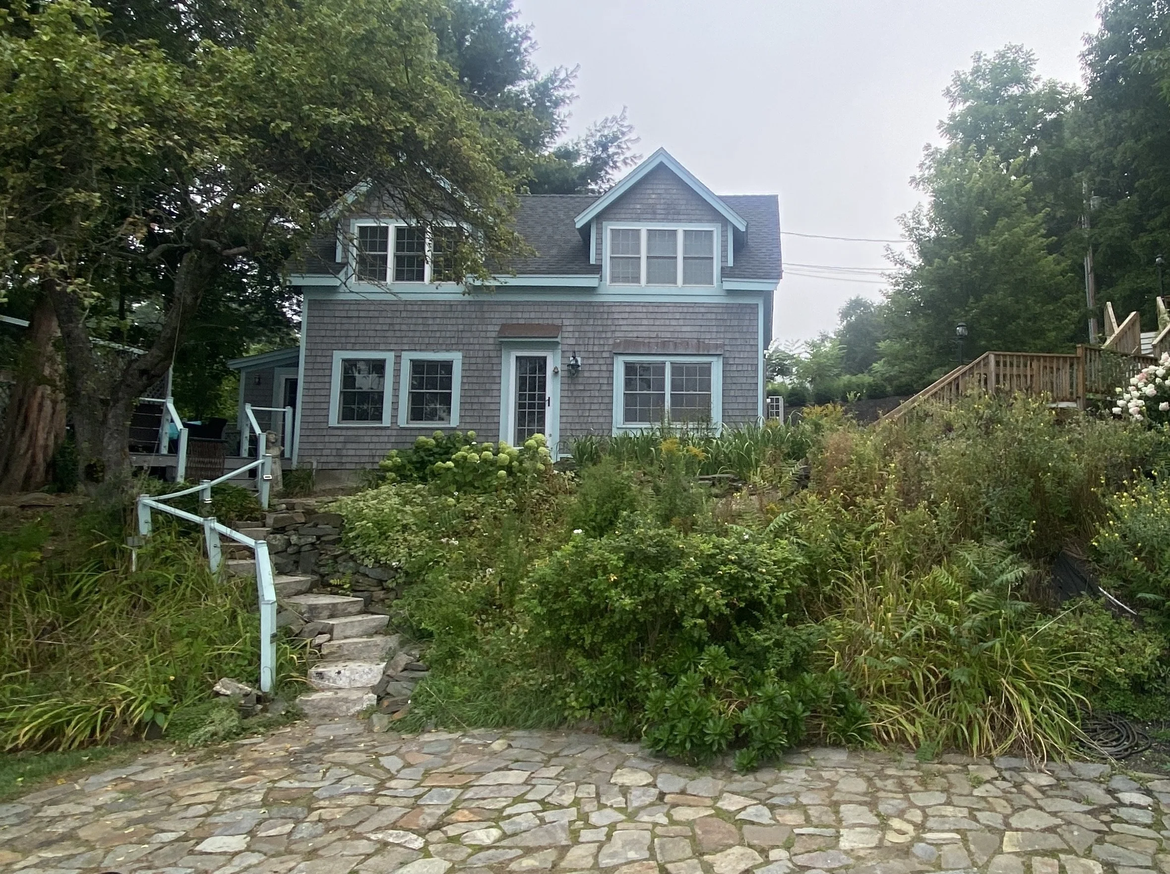 A two-story house with blue trim and gray shingles, set on a hill with stone steps leading up to the front door. The house has multiple windows and is surrounded by green vegetation and trees, with a stone pathway in the foreground.
