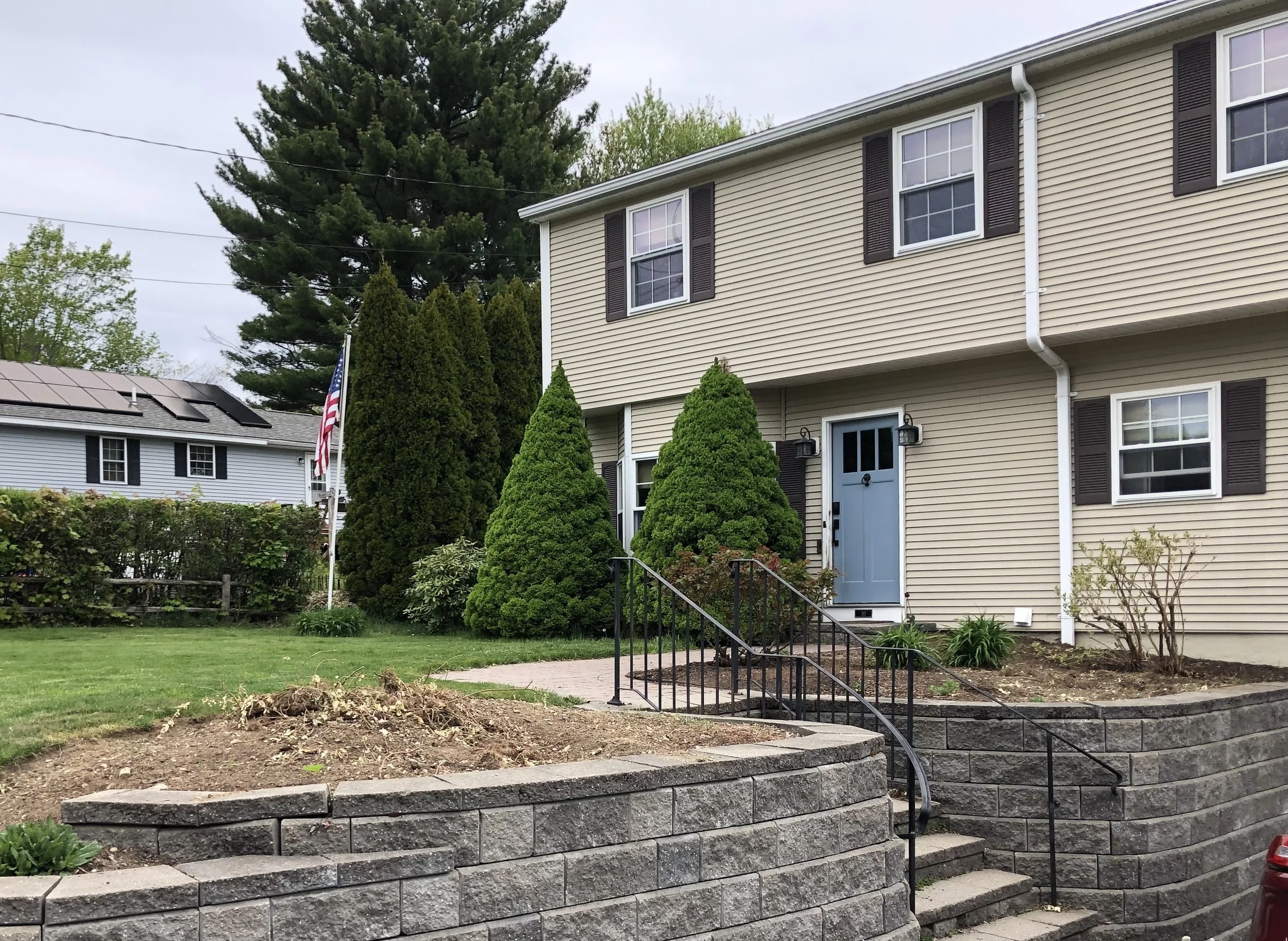 A two-story house with beige siding, dark brown shutters, and a light blue front door. In front, there is a landscaped yard with a stone retaining wall, stairs, small bushes, and tall trees in the background.