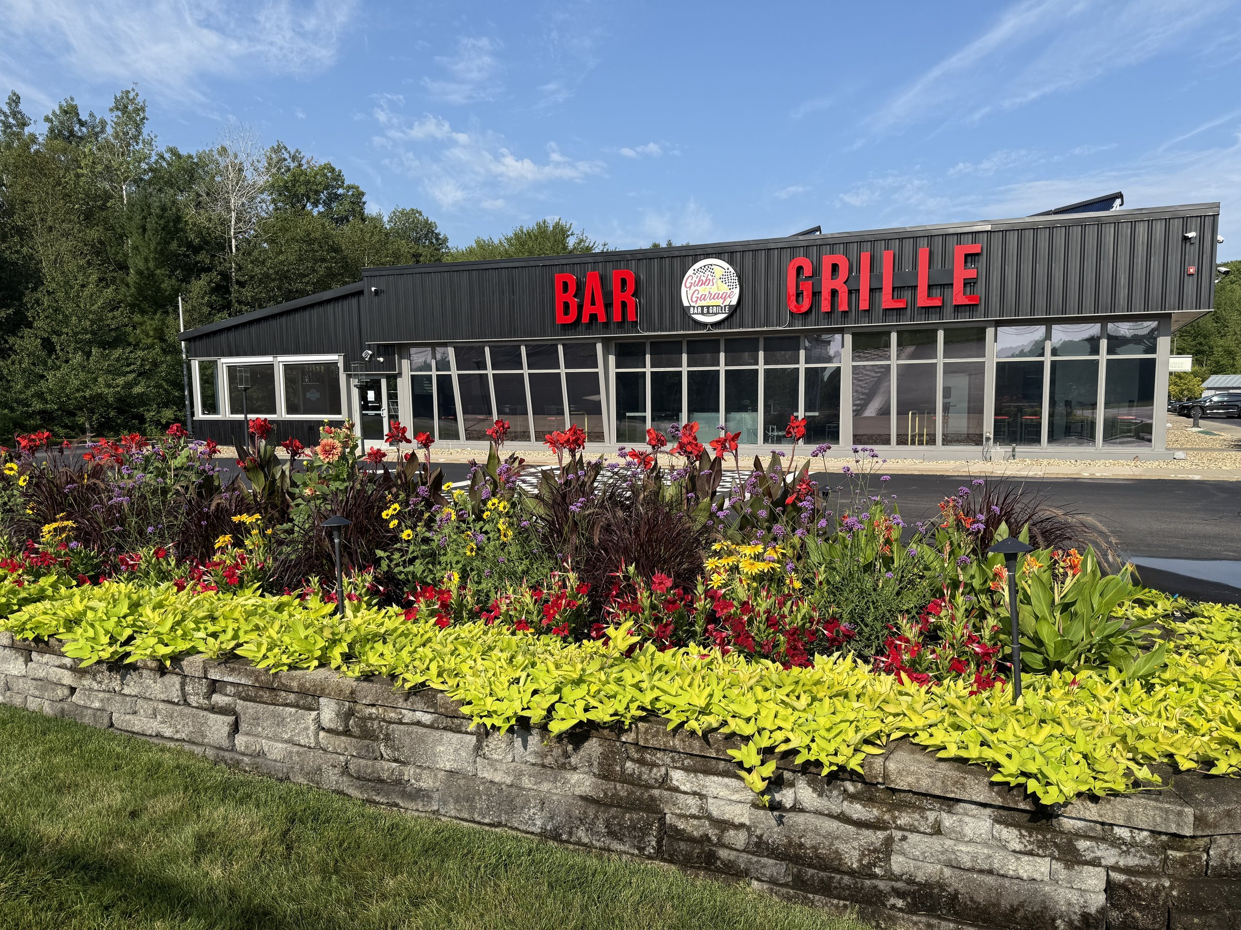 A building with a sign that reads "Bar" and "Grille" with a smaller sign in the middle that says "Gibbs Garage Bar & Grille". It is surrounded by a colorful flower garden and has a parking lot in front. There are trees and a blue sky in the backgroun
