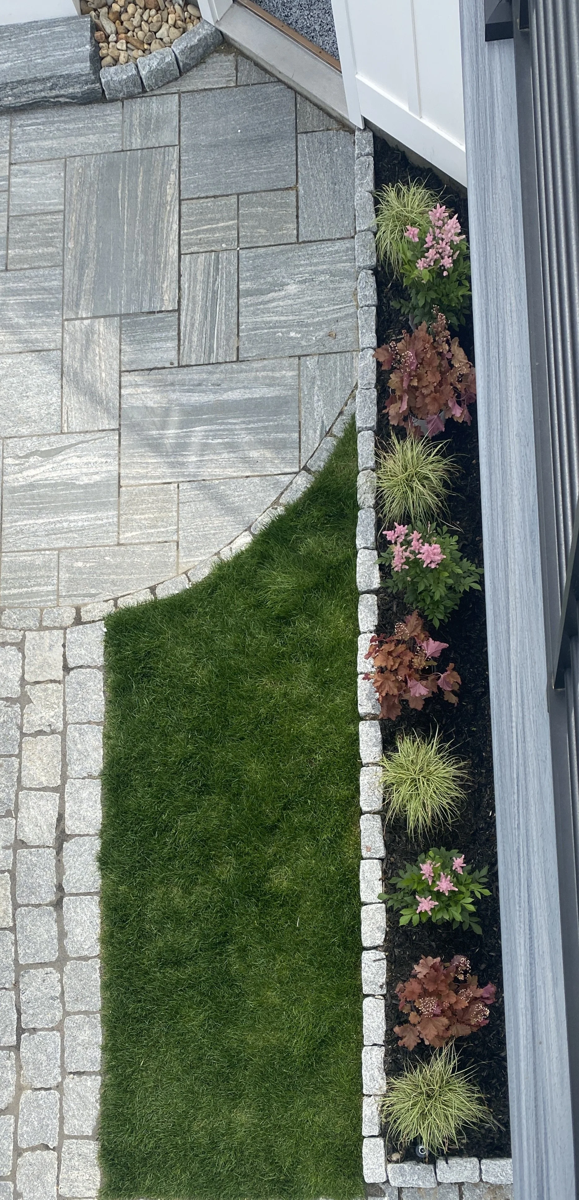 A landscaped garden with a stone pathway, green grass, and a flower bed with pink and reddish-brown plants bordered by gray bricks.