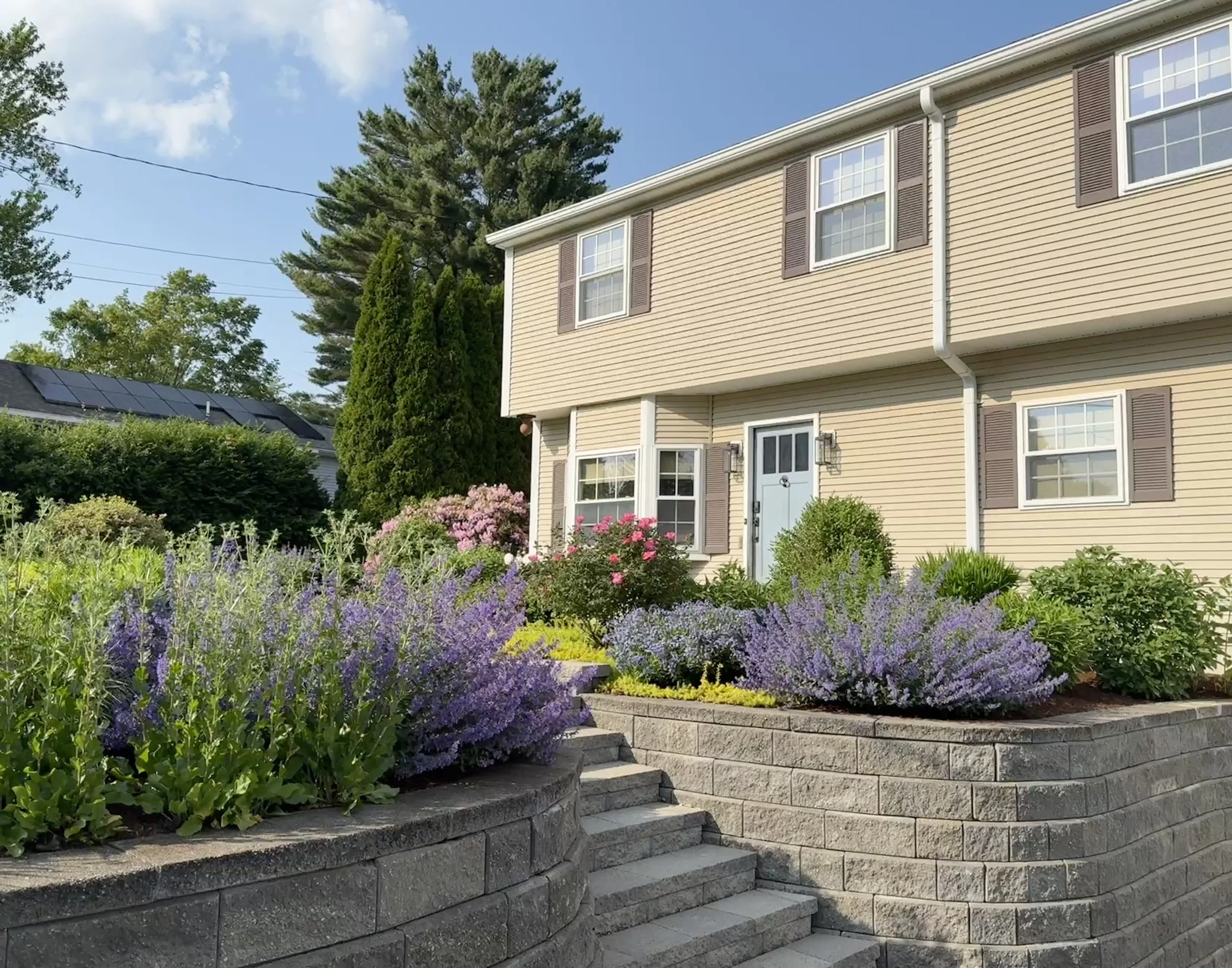 A beige two-story house with brown shutters and a light blue front door, surrounded by a well-maintained garden with purple and pink flowers, green bushes, and a stone retaining wall with steps, under a clear blue sky.