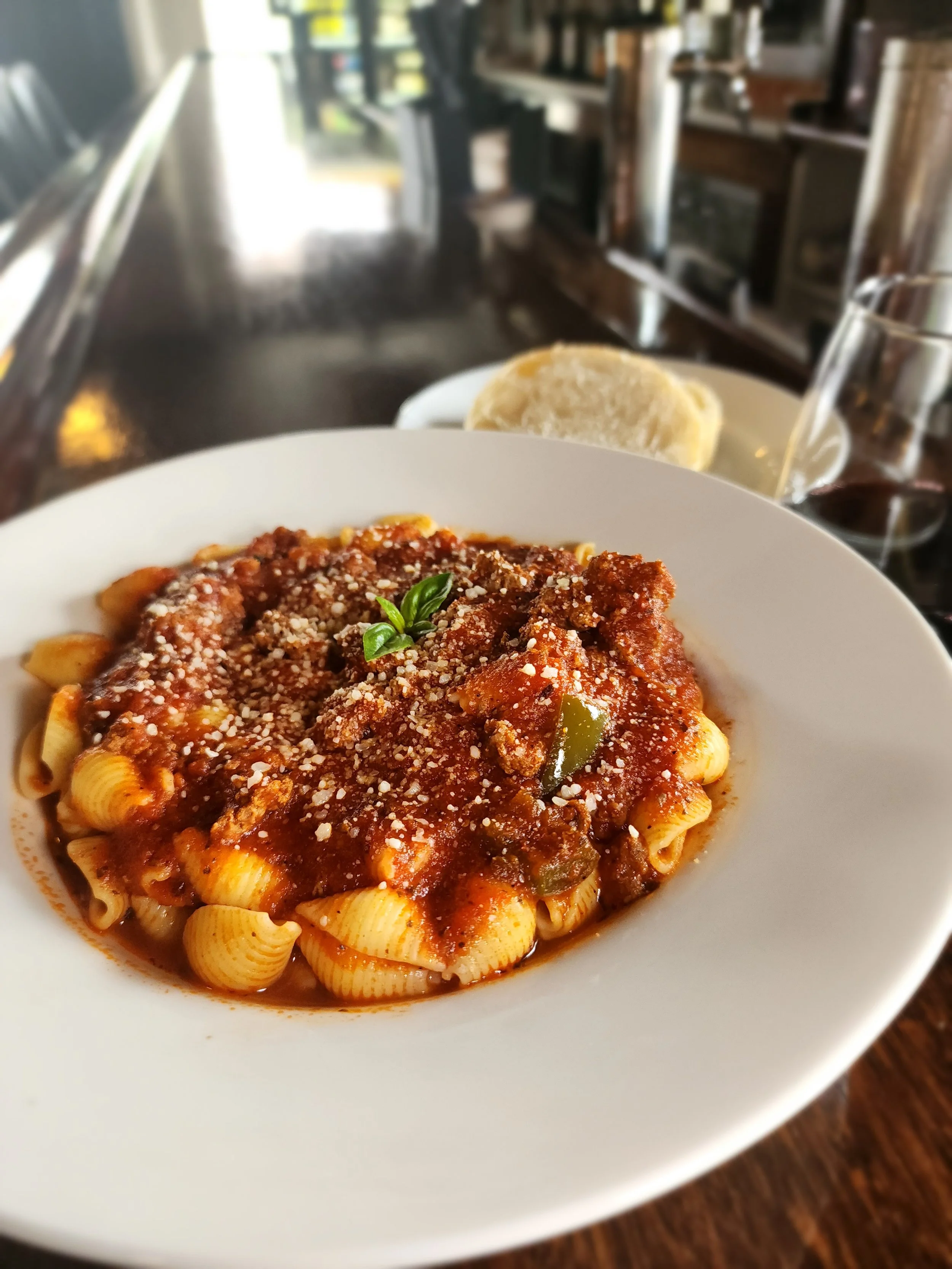 A bowl of homemade goulash with tomato sauce, garnished with grated cheese and a basil leaf, on a wooden table. In the background, a piece of bread and a glass of red wine are visible.