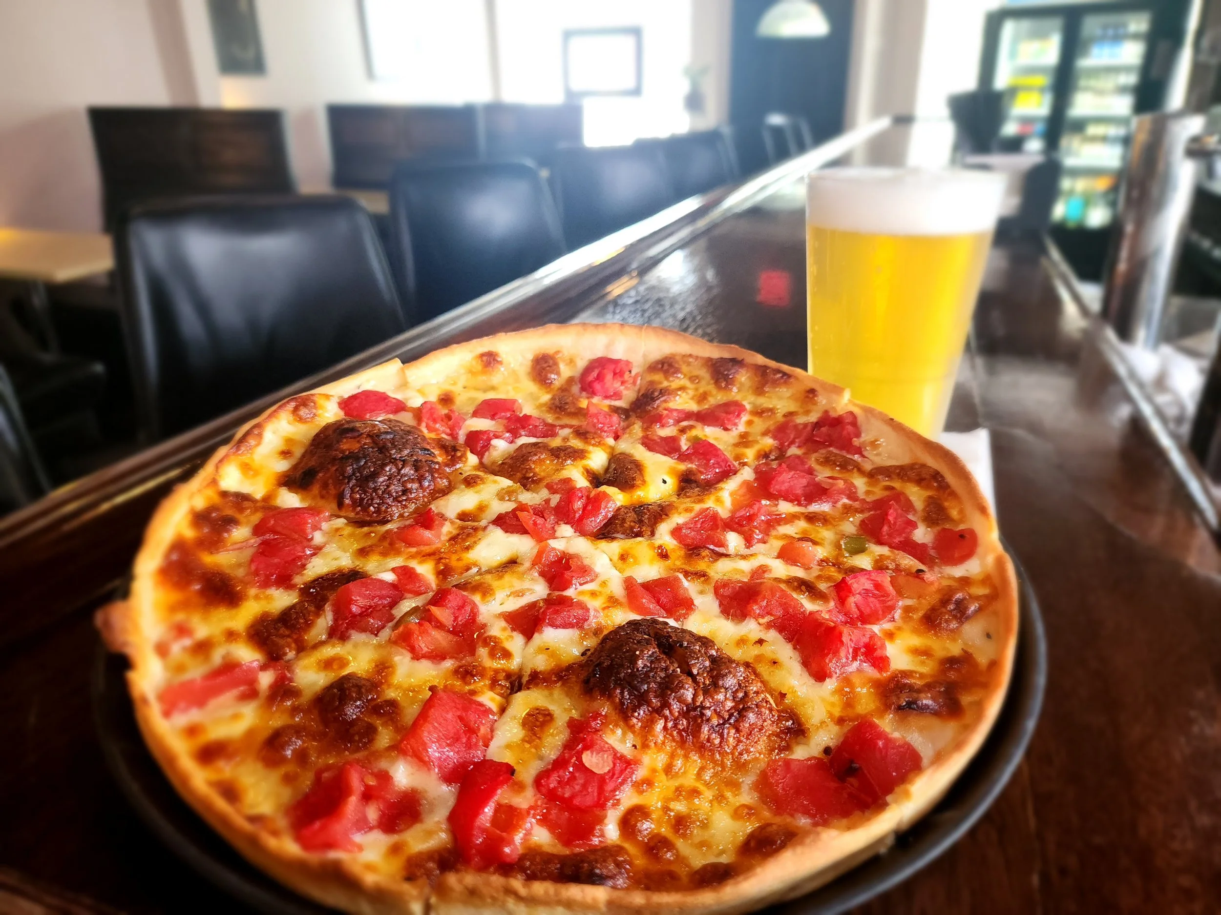 Nonni's oil and garlic pizza with fresh diced tomatoes on a black plate with a glass of beer on a wooden bar counter in a restaurant.