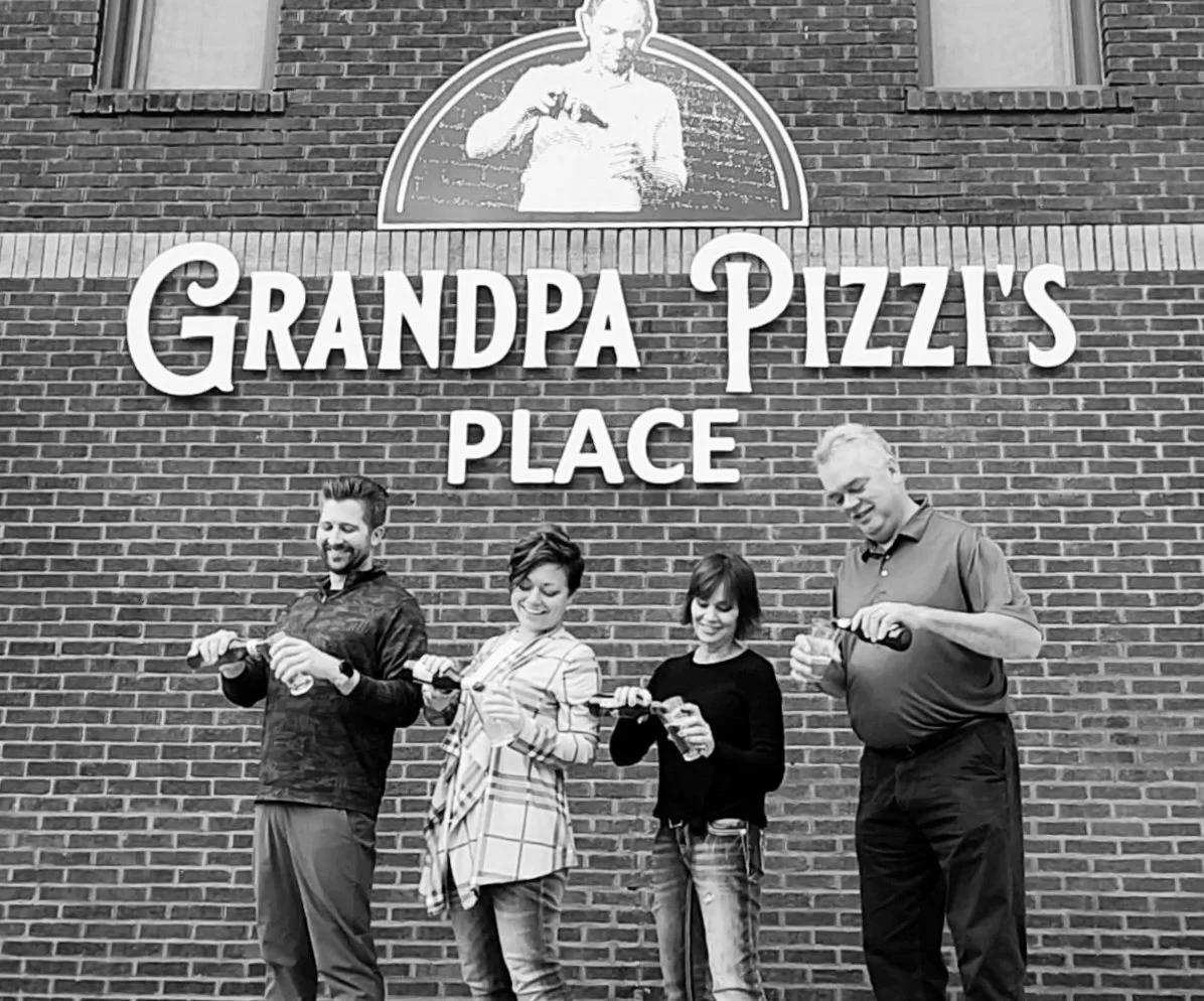 Group the current owners standing in front of a brick wall with a sign that reads "Grandpa Pizza's Place," opening bottles of drinks and smiling. Pictured from left to right: Alec Hill, Taylor Hill, Karen Wheeler, and Mark Wheeler.
