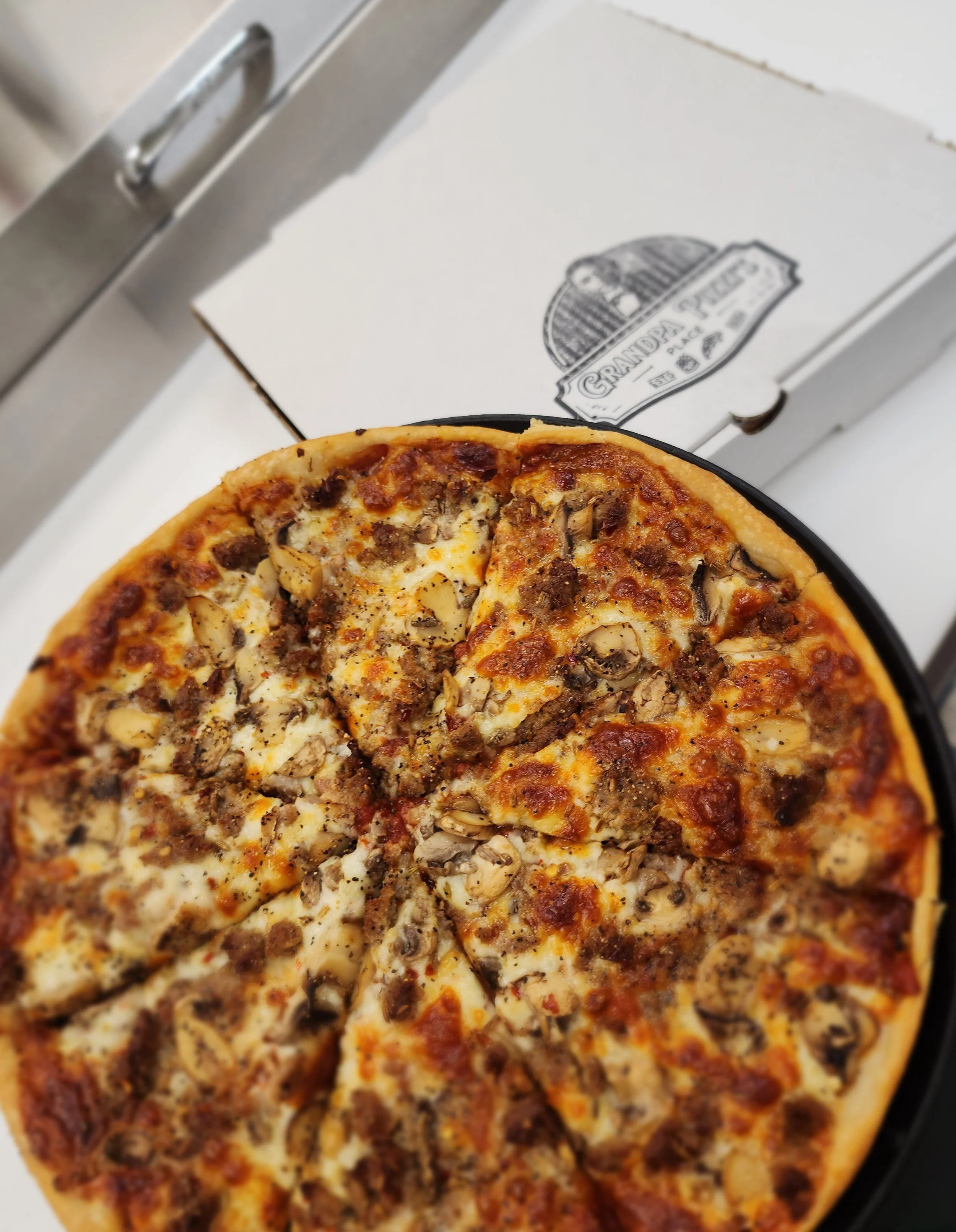 A specialty pizza with sausage, mushrooms, and cheese on a black plate, next to a white pizza box labeled 'Grandpa's'.