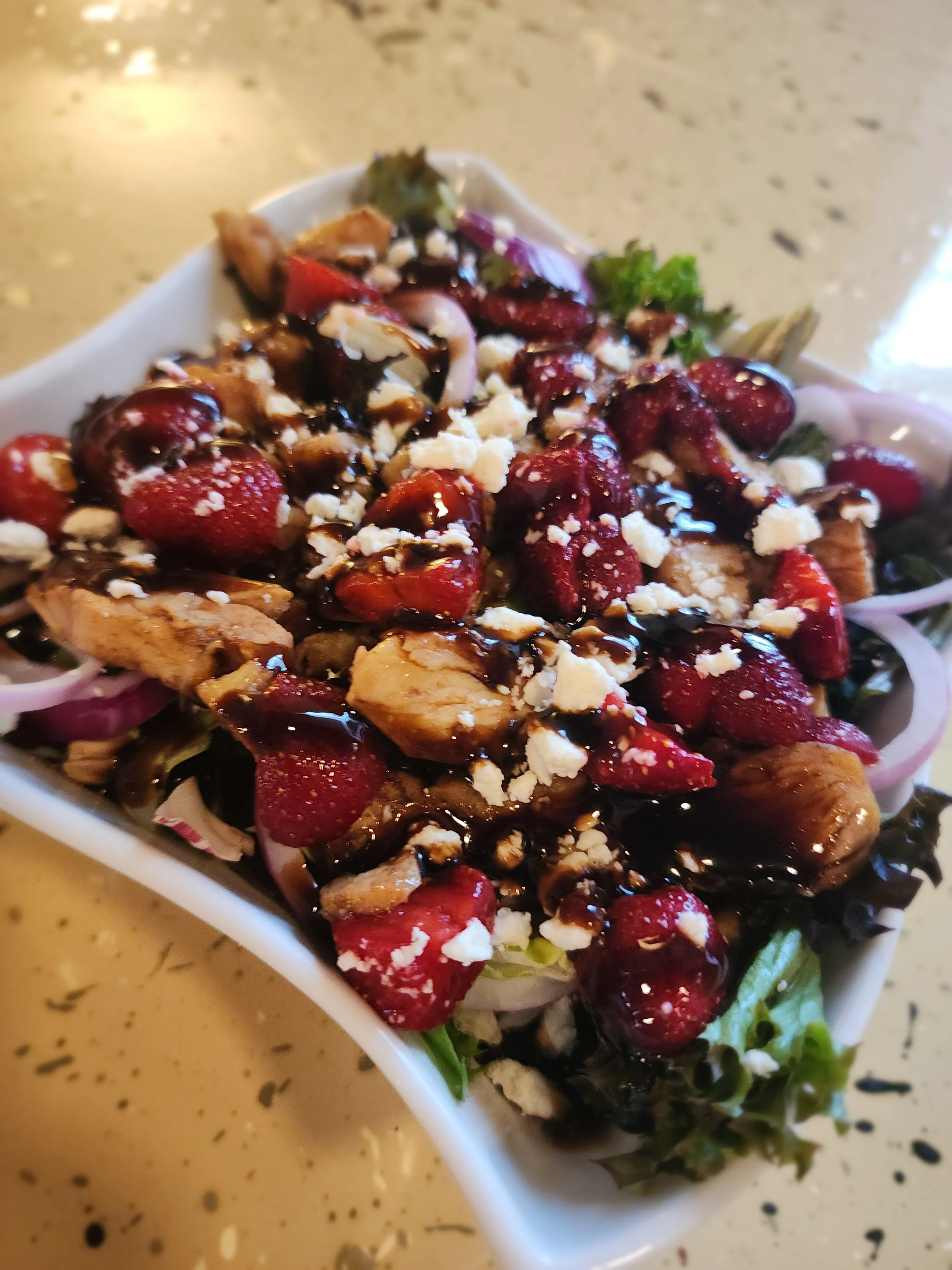 A salad with mixed greens, cranberries, grilled chicken, freshly grated cheese, and balsamic glaze in a white bowl.