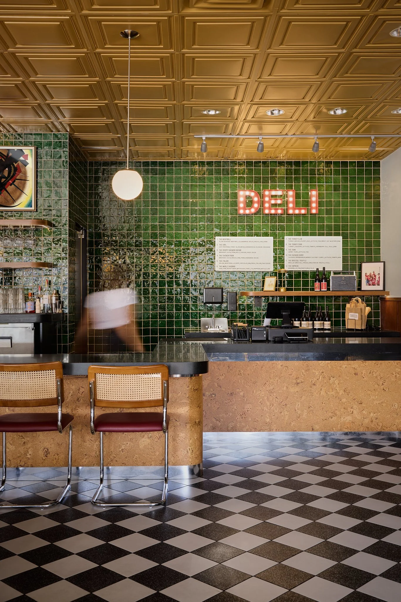 Mid-century modern hospitality project in Encinitas featuring cork wrapped bar and deli counter with chrome accents, vintage pendant lighting, mustard colored ceiling tiles, black and white checkered terrazzo floor tile, deep green wall tiles and cus