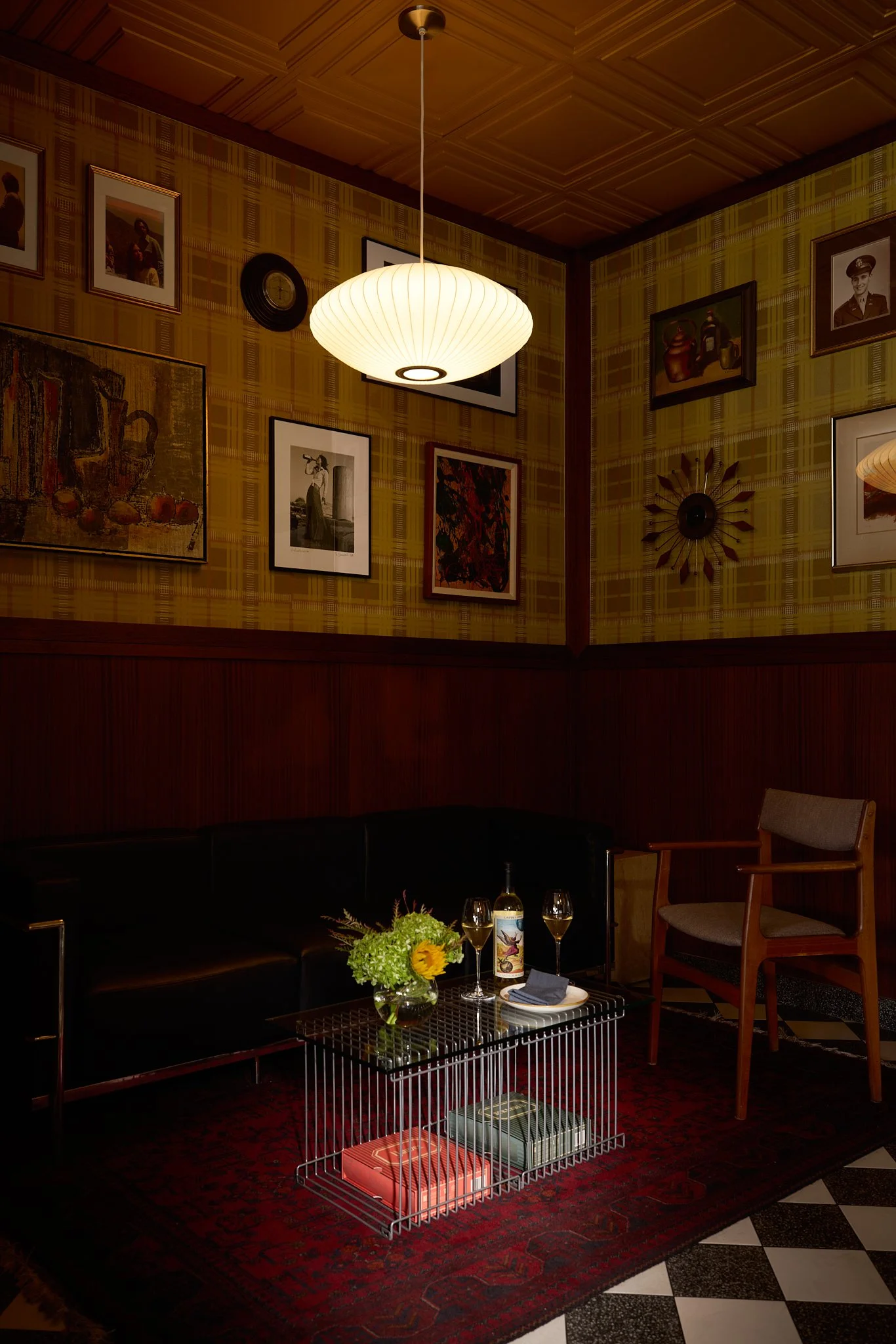 Mid-century modern hospitality project lounge area featuring vintage black leather sofa, vintage chairs, chrome and glass coffee table, wood wall paneling, George Nelson pendant light, plaid wallpaper and gallery wall of vintage art and family photos
