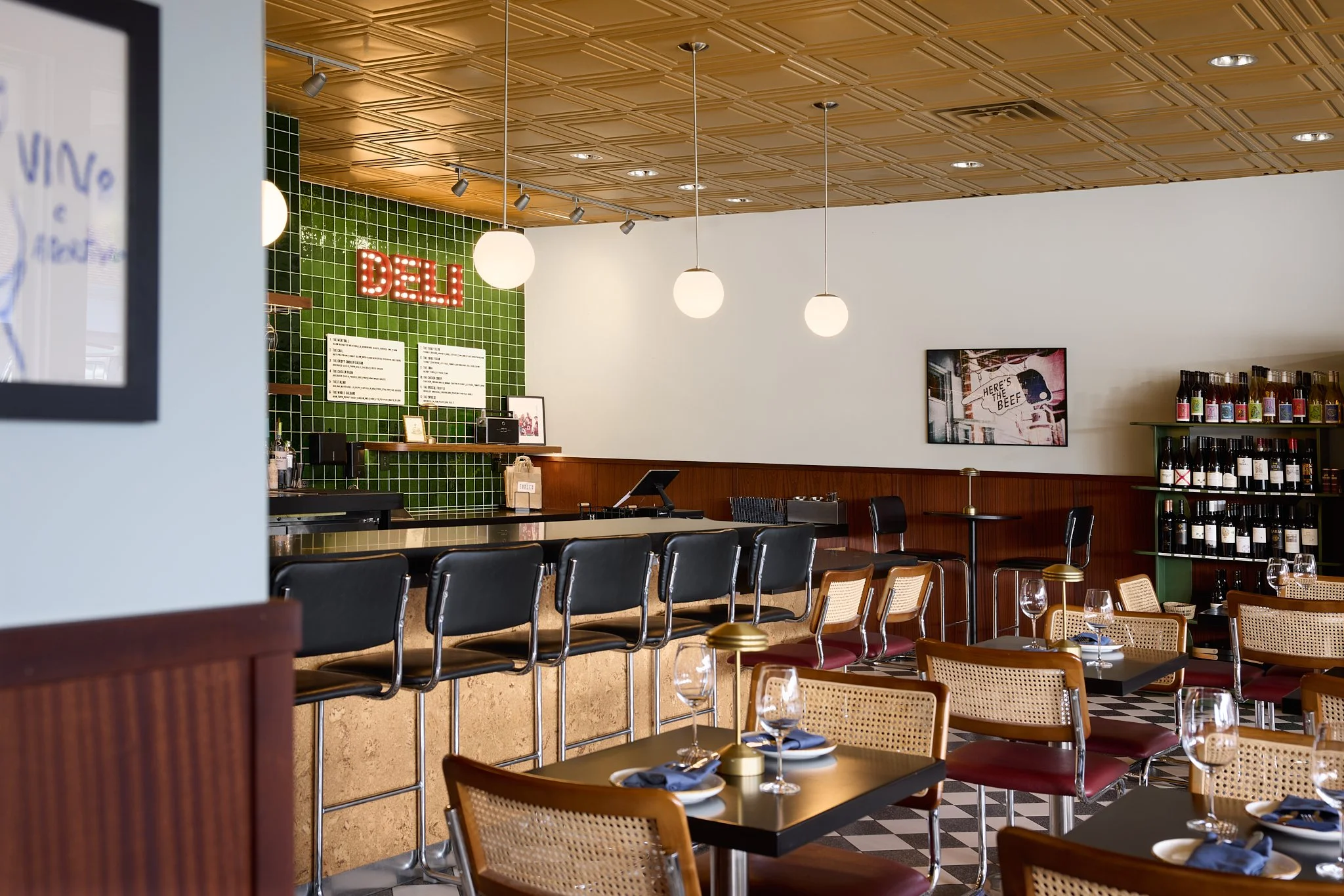 Mid-century modern restaurant design featuring black and white terrazzo checkered floor tiles, cork bar with chrome accents, wood paneling, rich burgundy seating, and deep green wall tiles. 