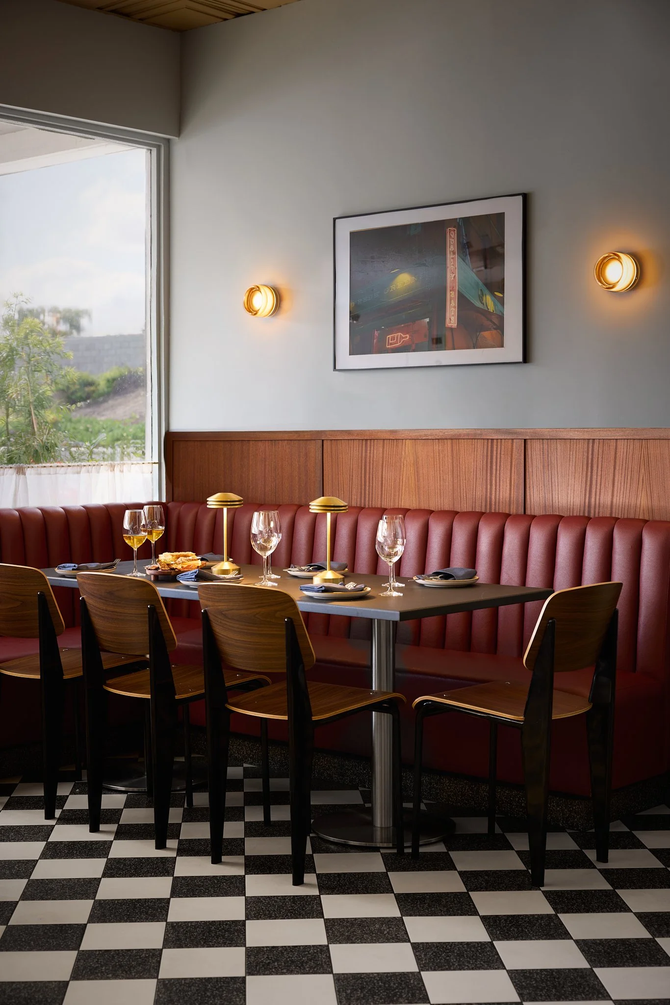 Mid-century modern restaurant and wine bar design in Encinitas featuring rich burgundy banquette seating, cafe curtains, vintage style walnut seating and wood wall paneling, checkered terrazzo floor tiles, art, and modern wall sconces.