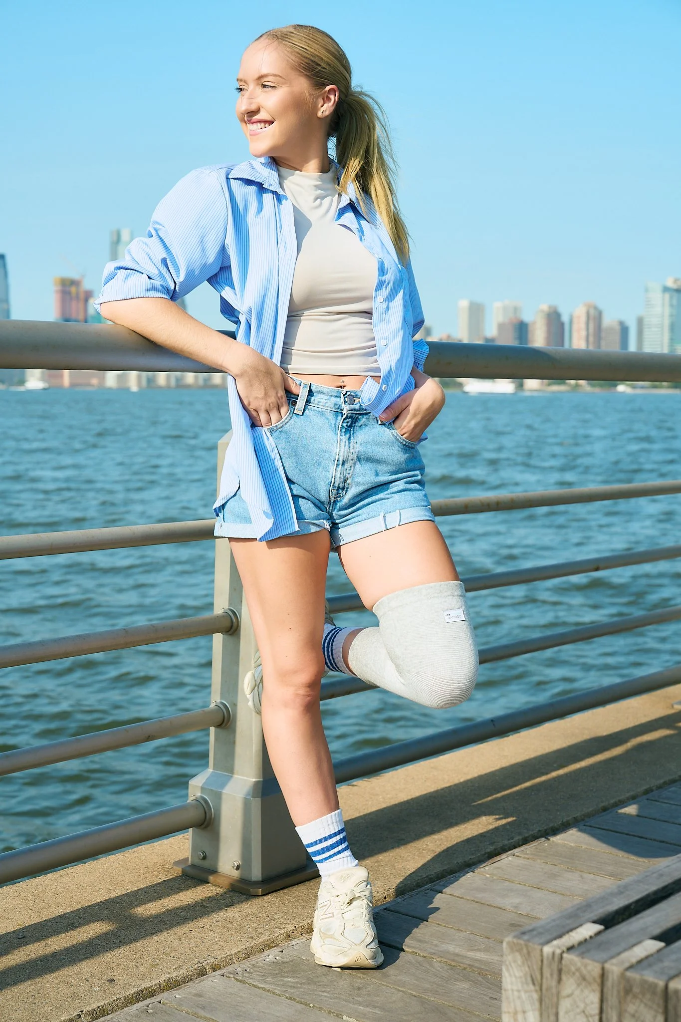 Young woman with blonde hair in a ponytail smiling, leaning against a railing by the water, wearing a blue striped shirt, white t-shirt, denim shorts, and white sneakers, with a city skyline in the background.