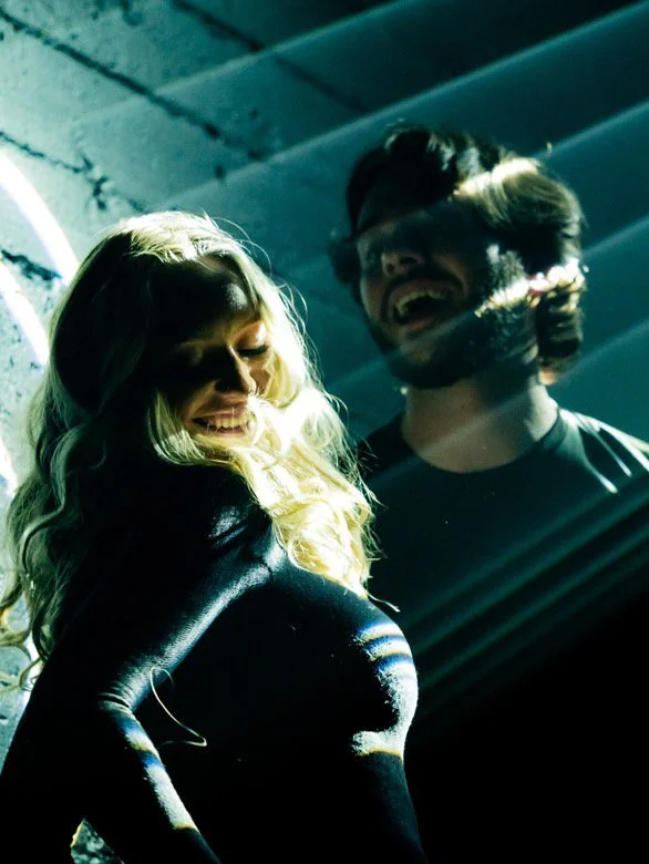 Two people, a woman with long blonde hair and a man with sunglasses and facial hair, laughing together in a dimly lit environment.
