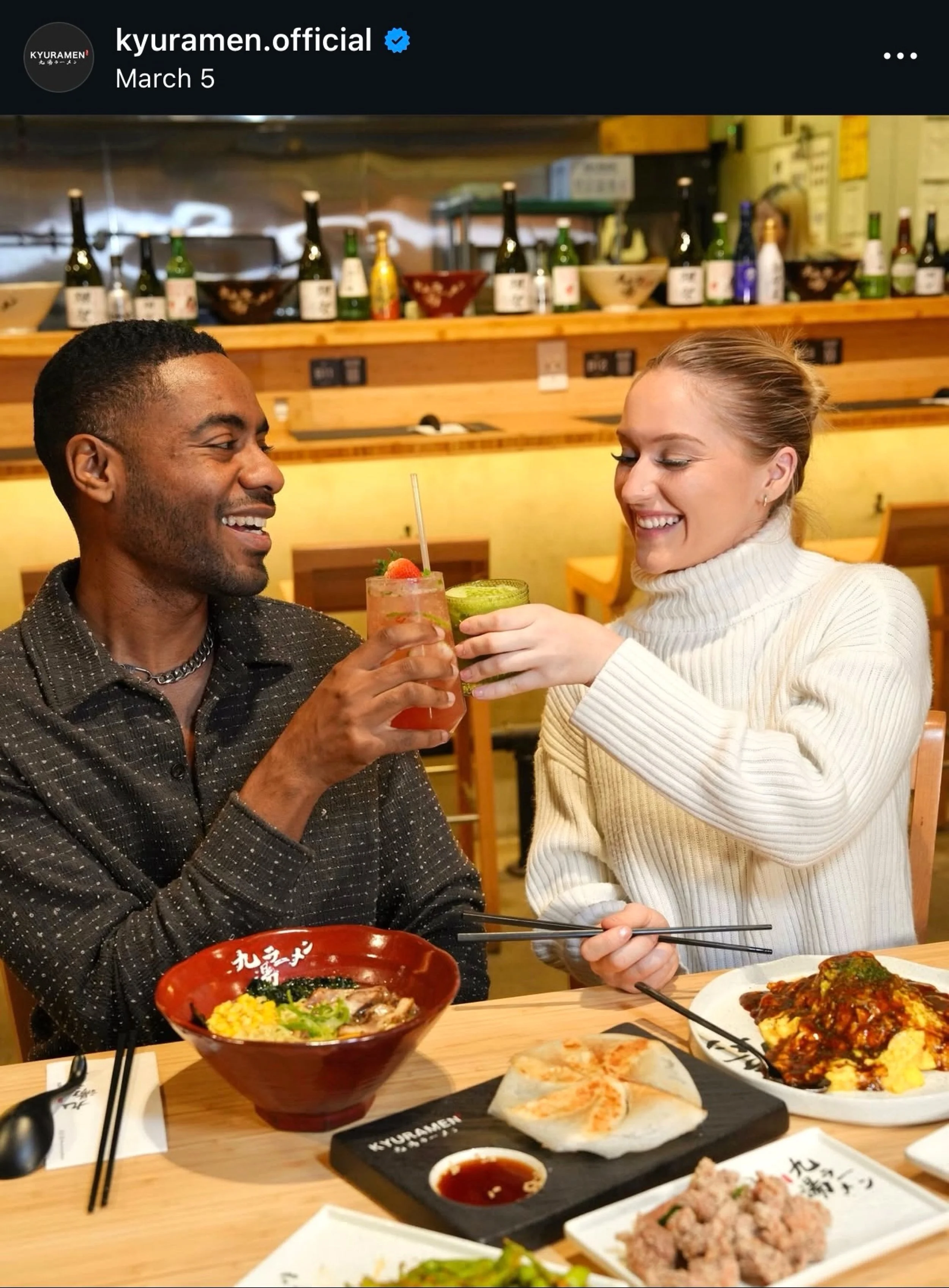 A man and a woman sitting at a restaurant table, smiling and clinking drinks. The man is holding a cocktail with a cherry and straw, while the woman has a green drink. The table has various dishes including ramen, a dish with sauce and rice, and othe
