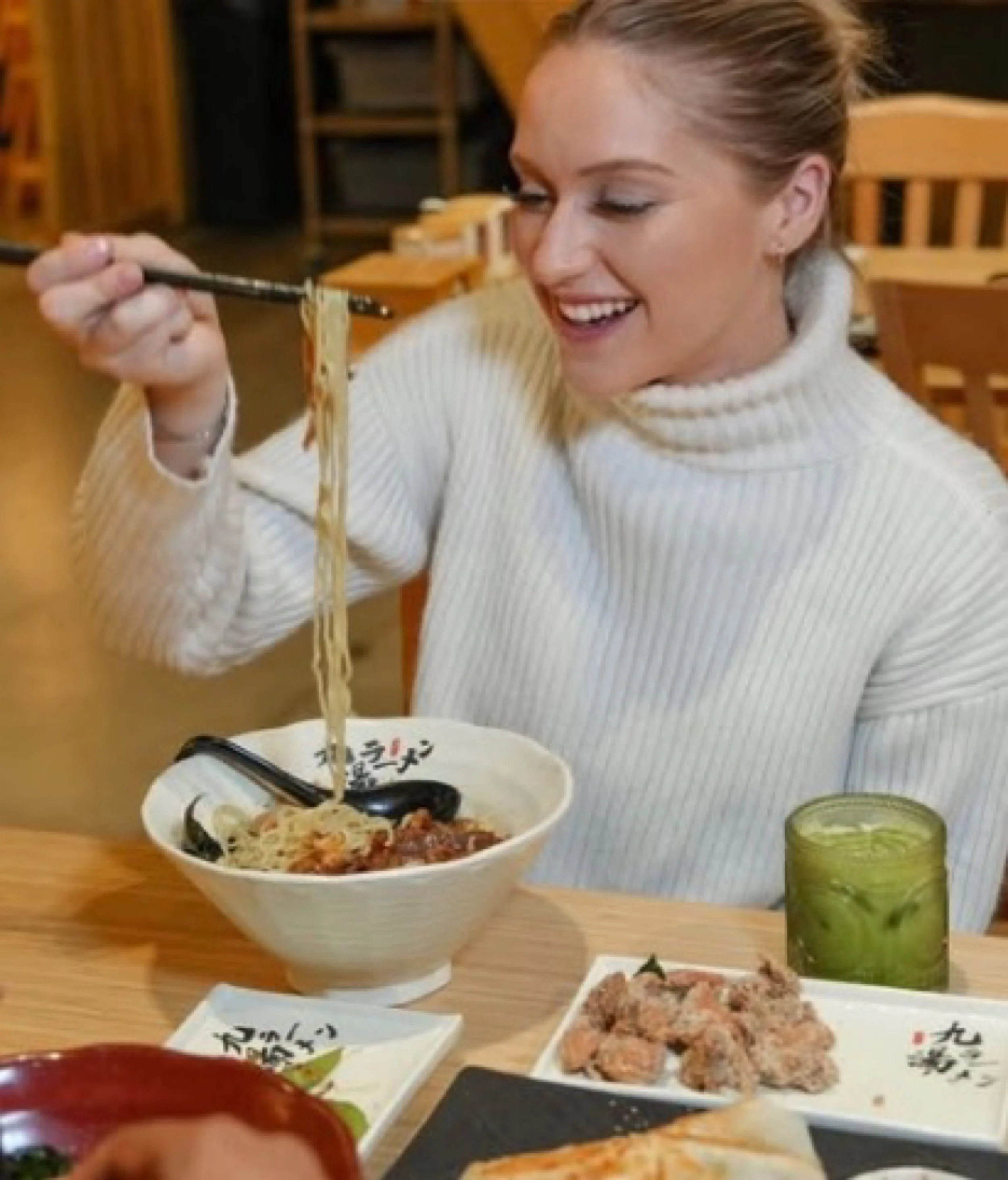 A woman with blonde hair in a white sweater sitting at a wooden table in a restaurant, lifting noodles from a bowl of ramen with chopsticks, smiling.