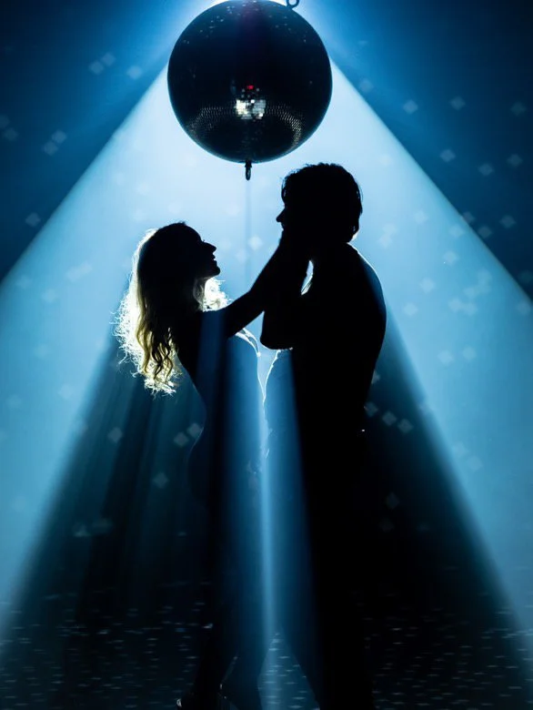 Silhouettes of a man and woman dancing under a shiny disco ball in a dimly lit club with beams of light.