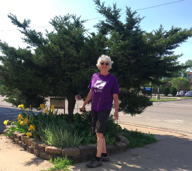 An elderly woman with white hair and sunglasses standing outdoors next to a landscaped flower bed with yellow flowers and greenery, in front of a large tree, on a sidewalk near a street.
