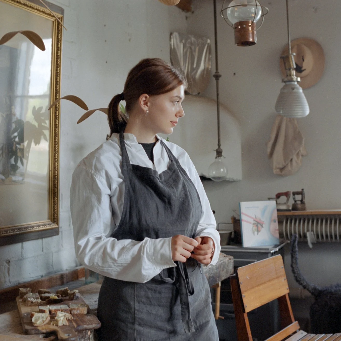 A woman in a black apron and white shirt standing in a cozy room with vintage decor, looking to her left. There are light bulbs hanging from the ceiling, a mirror on the wall, and a wooden table with assorted food items.