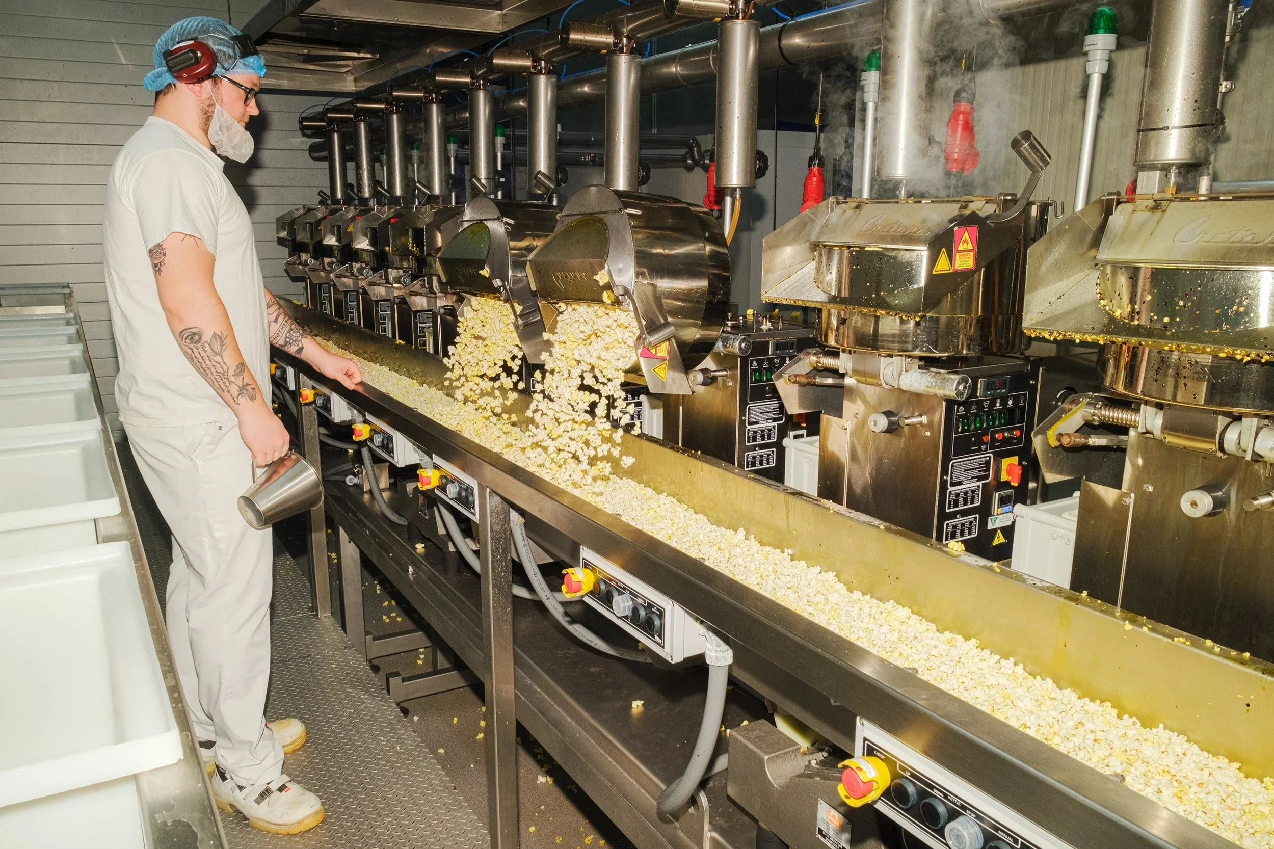 En person i operationströja och skyddsglasögon använder maskin som poppar popcorn i en fabrik.
