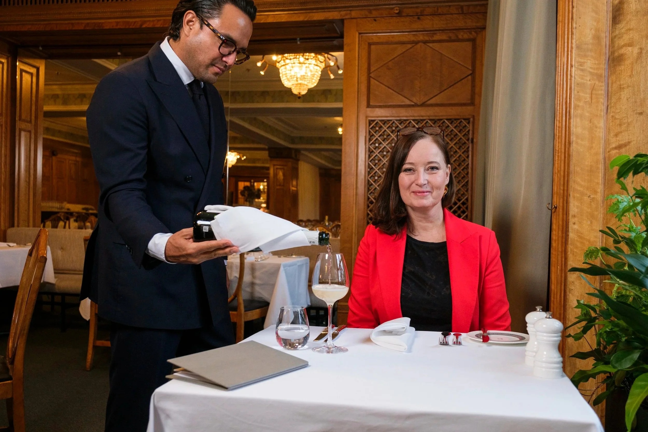 En man i mörk kostym serverar vin till en kvinna i röd kavaj som sitter vid ett restaurangbord.