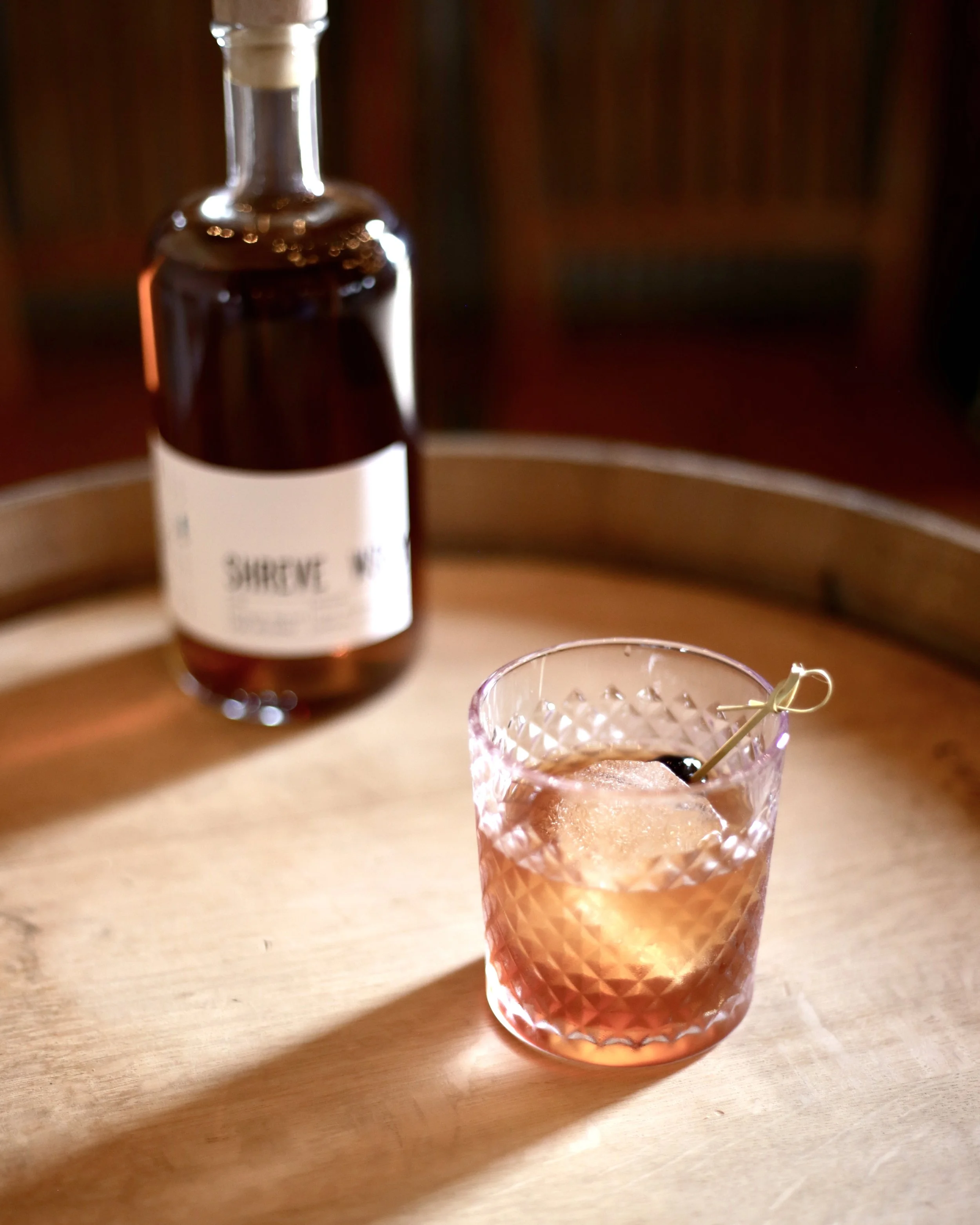 A close-up of a textured glass filled with a cocktail and an ice cube, with a toothpick garnish. In the background, there is a bottle of whiskey with a white label on a wooden surface.
