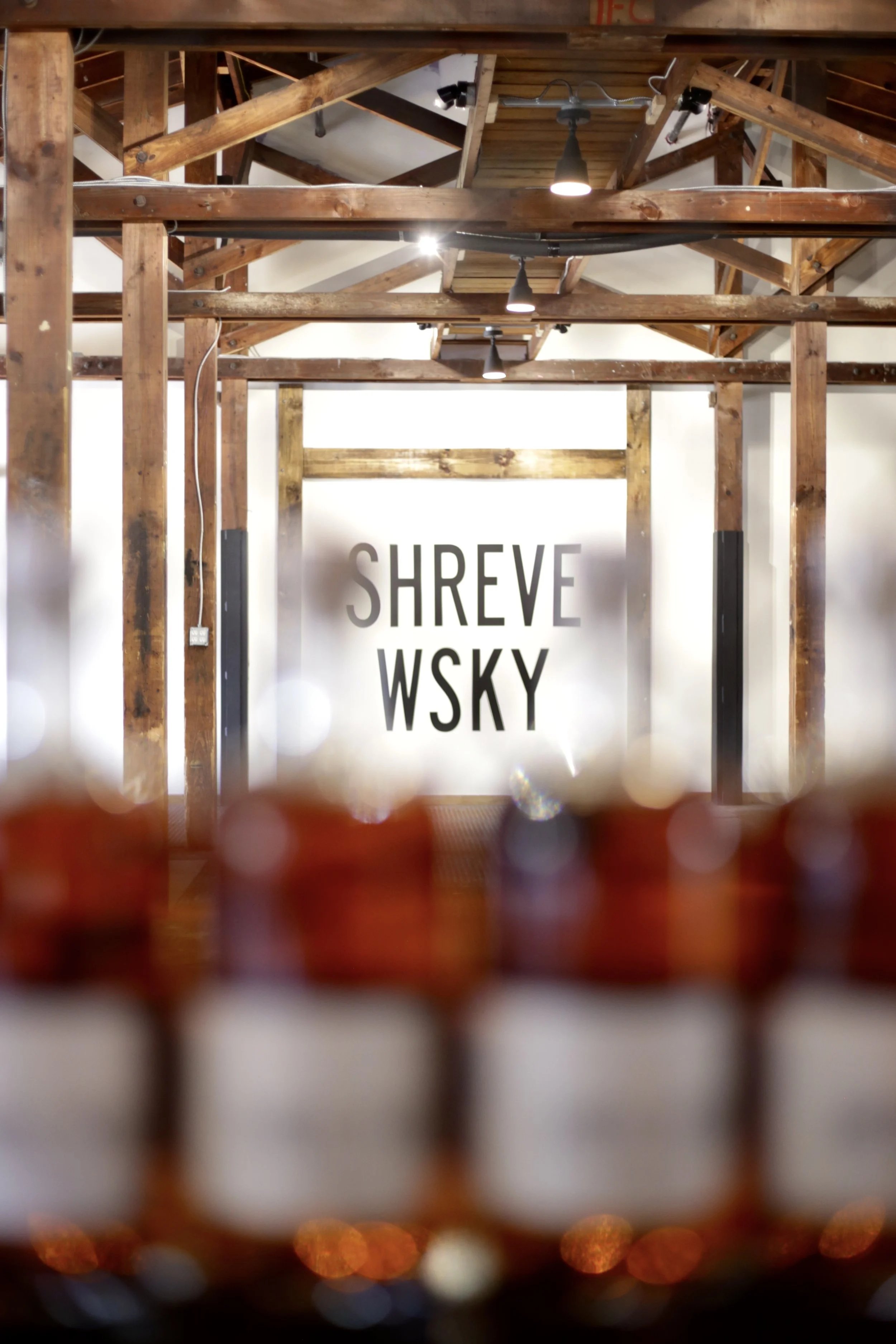 View through a row of whiskey bottles at a bar with an off-white wall in the background. Painted on a the wall is a sign that reads 'SHREVE WSKY'.