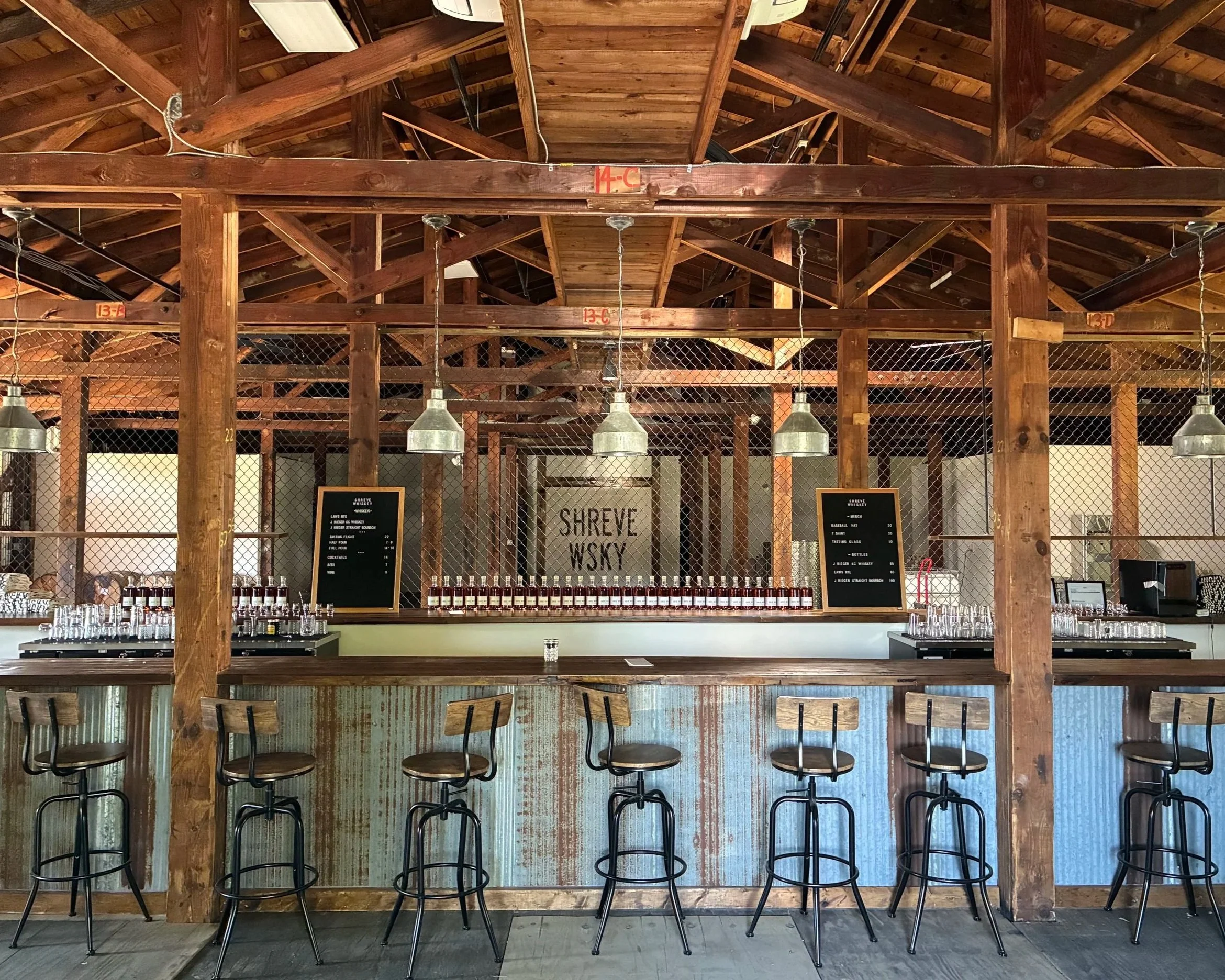 The interior of Shreve Whiskey, a rustic bar and whiskey tasting room with wooden beams and furniture. There is a bar counter with bottles and glasses, and ceiling lights hanging down. Some equipment and barrels are visible behind a chain-link fence.