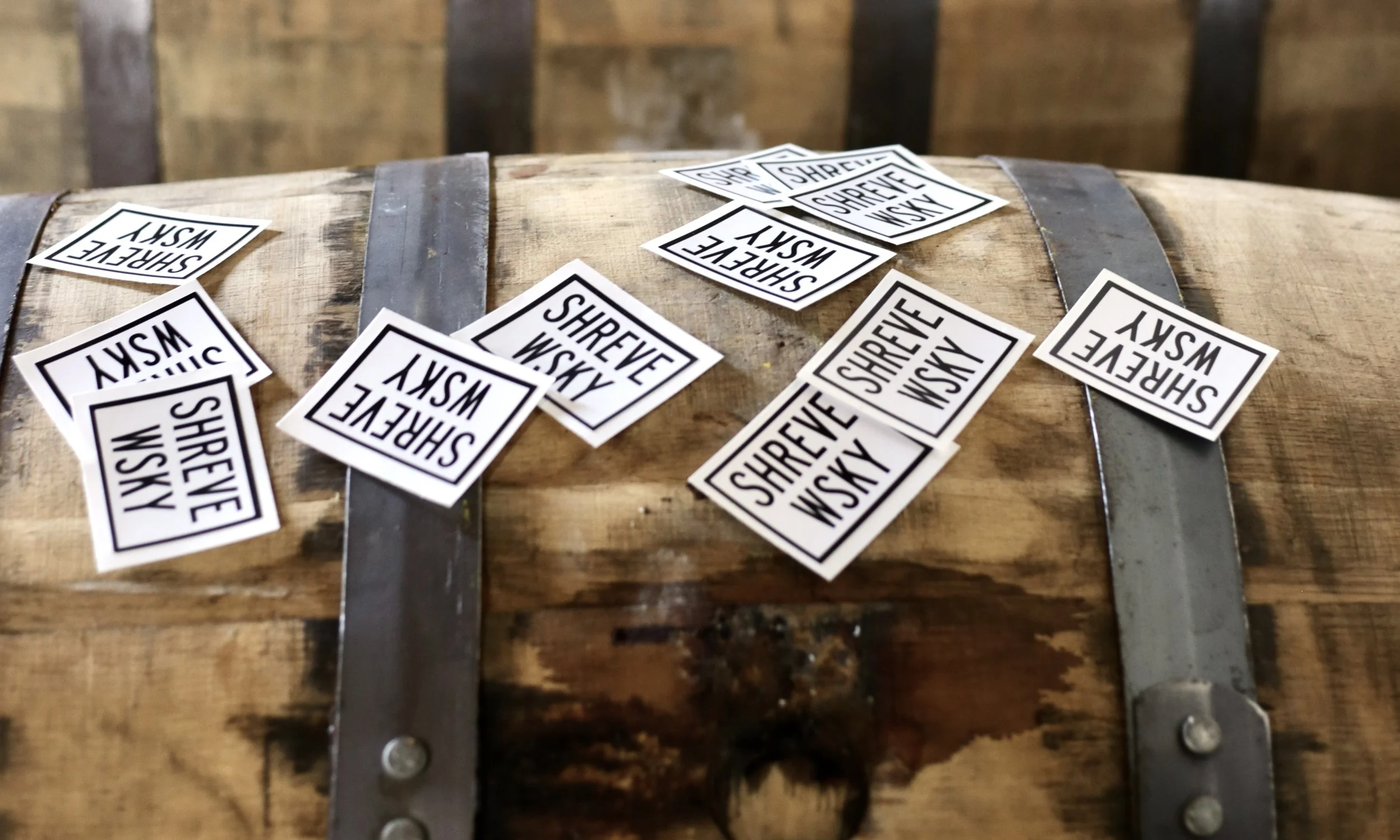 A wooden barrel with metal hoops, covered with white stickers that say 'SHREVE WSKY' and 'SHREVE WSKY' in black text, scattered on and around it.
