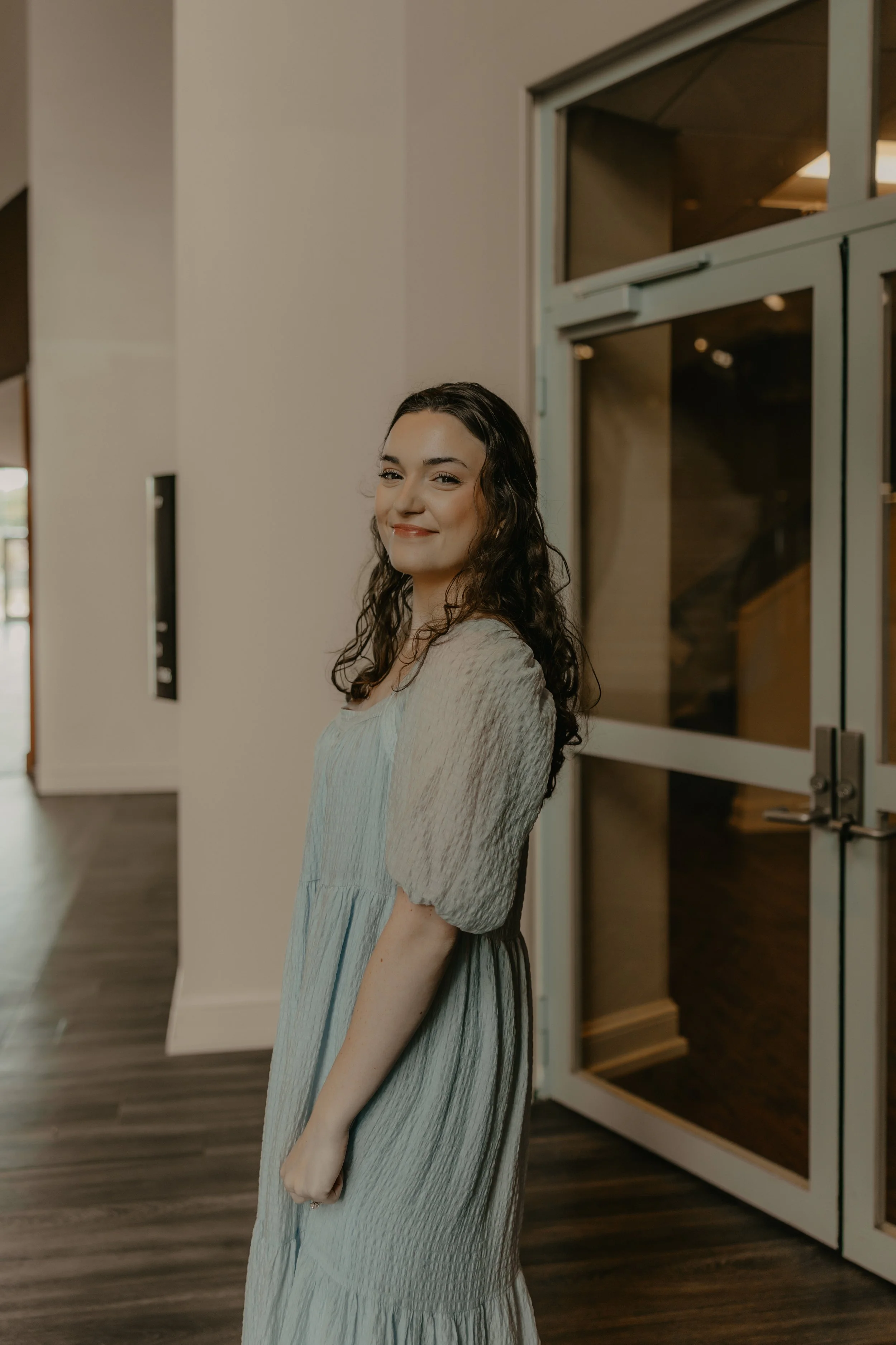 A young woman with long, wavy brown hair smiling and holding a professional camera with a large lens, standing inside a well-lit building with wooden floors and large windows.