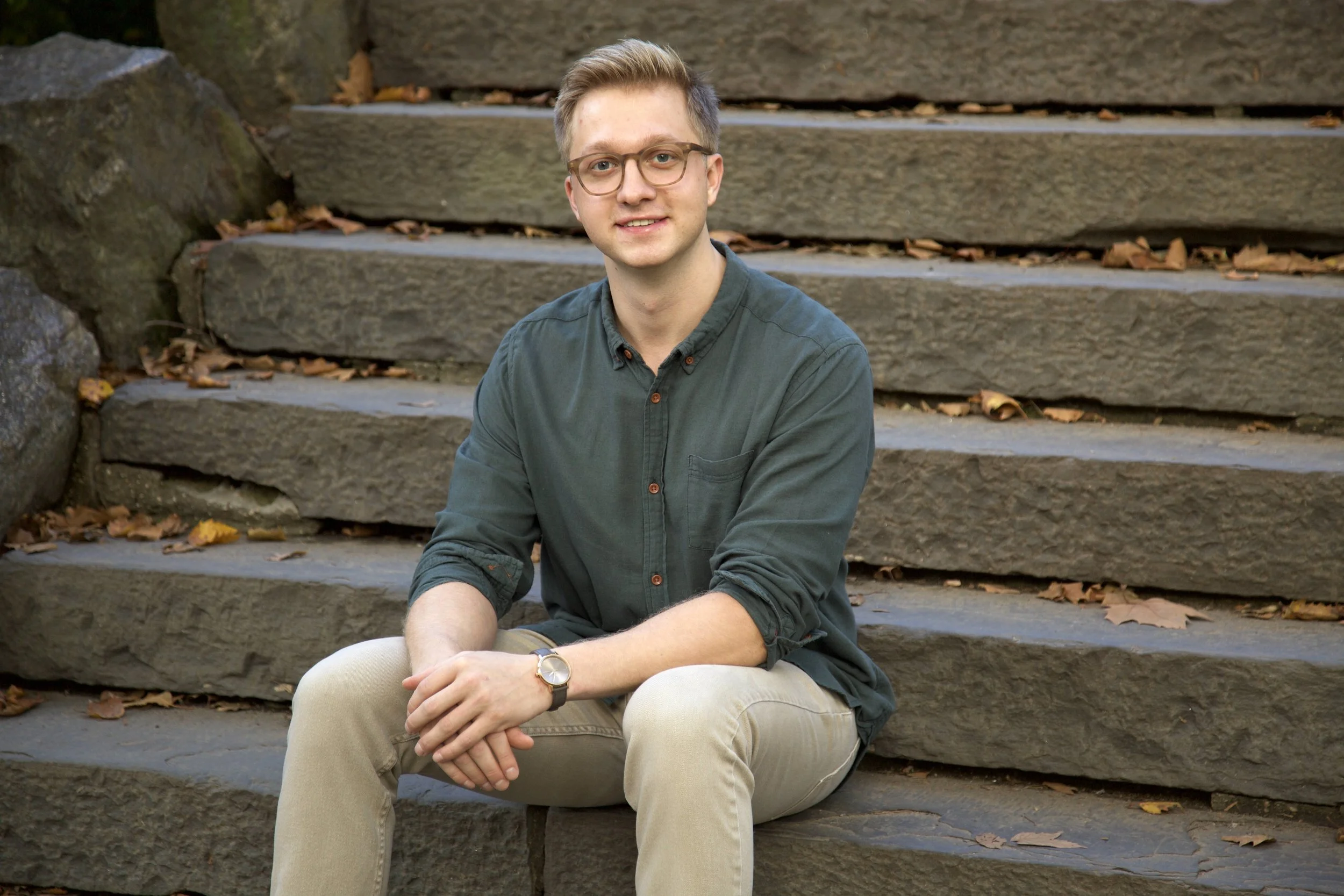 A young man with glasses and short blond hair sitting on outdoor stone steps, wearing a green button-up shirt and beige pants, with fallen leaves around him.