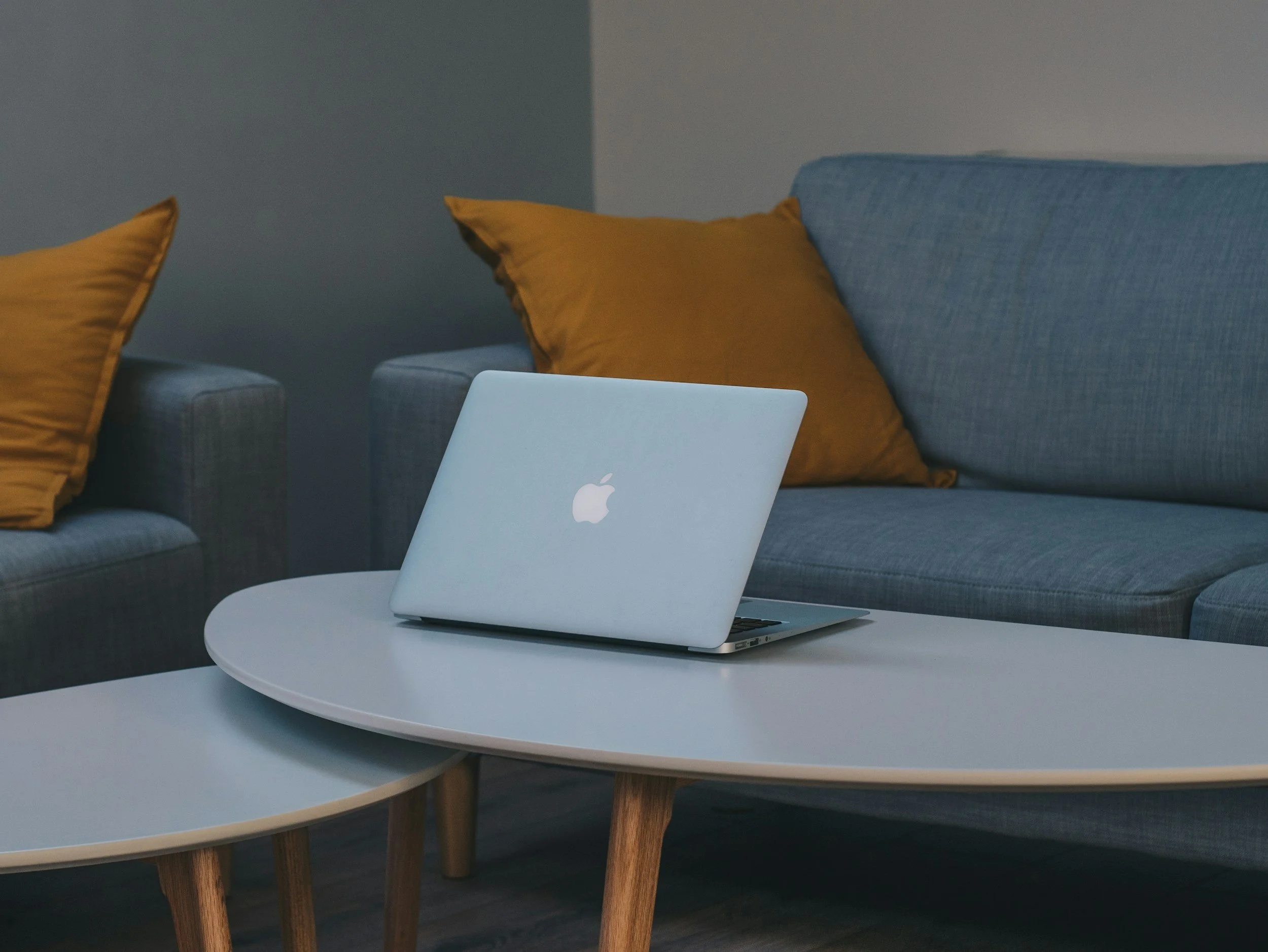A closed silver MacBook on a white round coffee table in a living room with gray sofa and mustard-colored pillows.