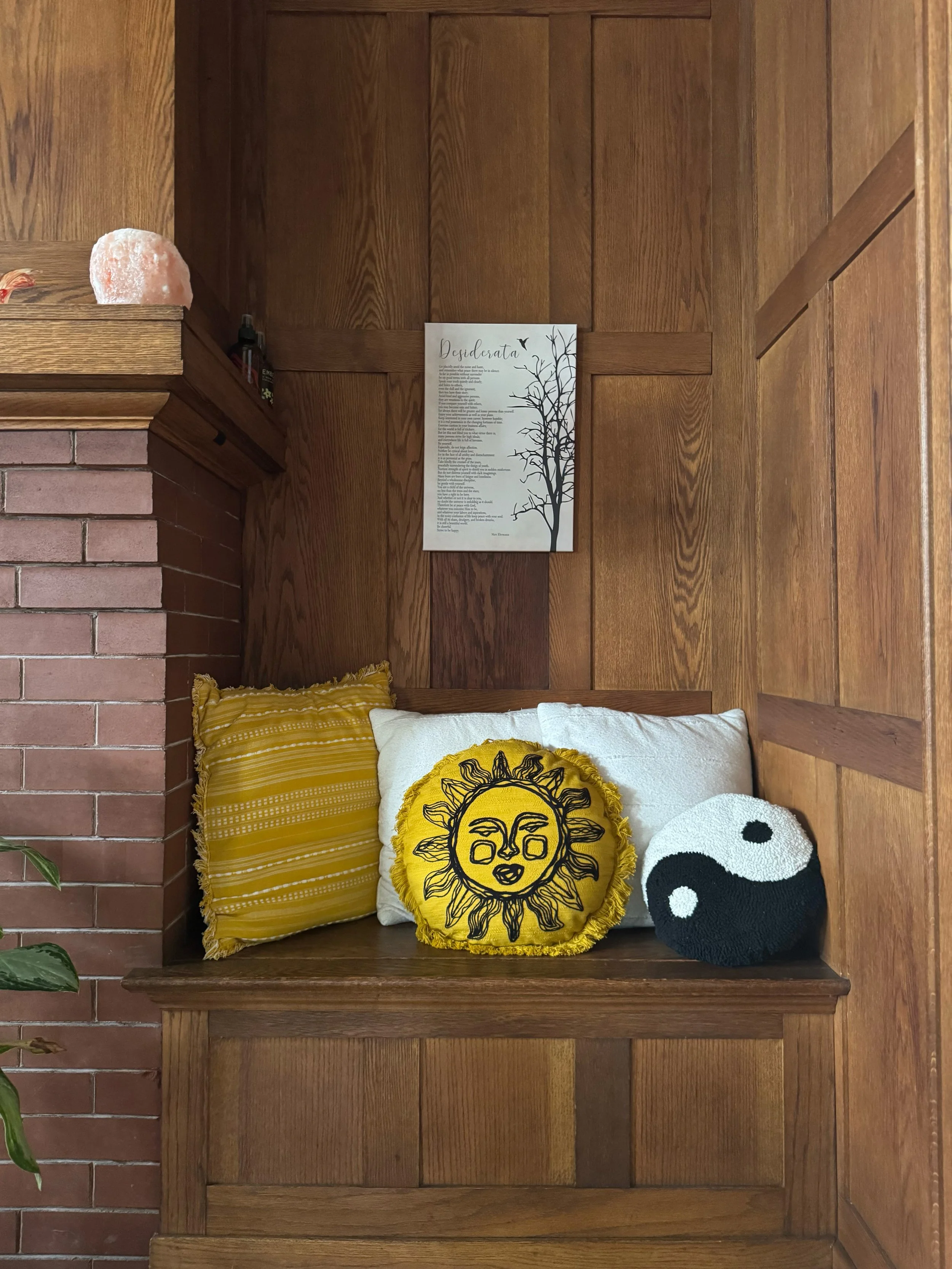 Wood-paneled nook with three decorative pillows: one yellow with horizontal stripes, one white with a black embroidered sun, and one black and white yin-yang. A brick wall is on the left, with a pink Himalayan salt lamp on the mantel. A framed poem o