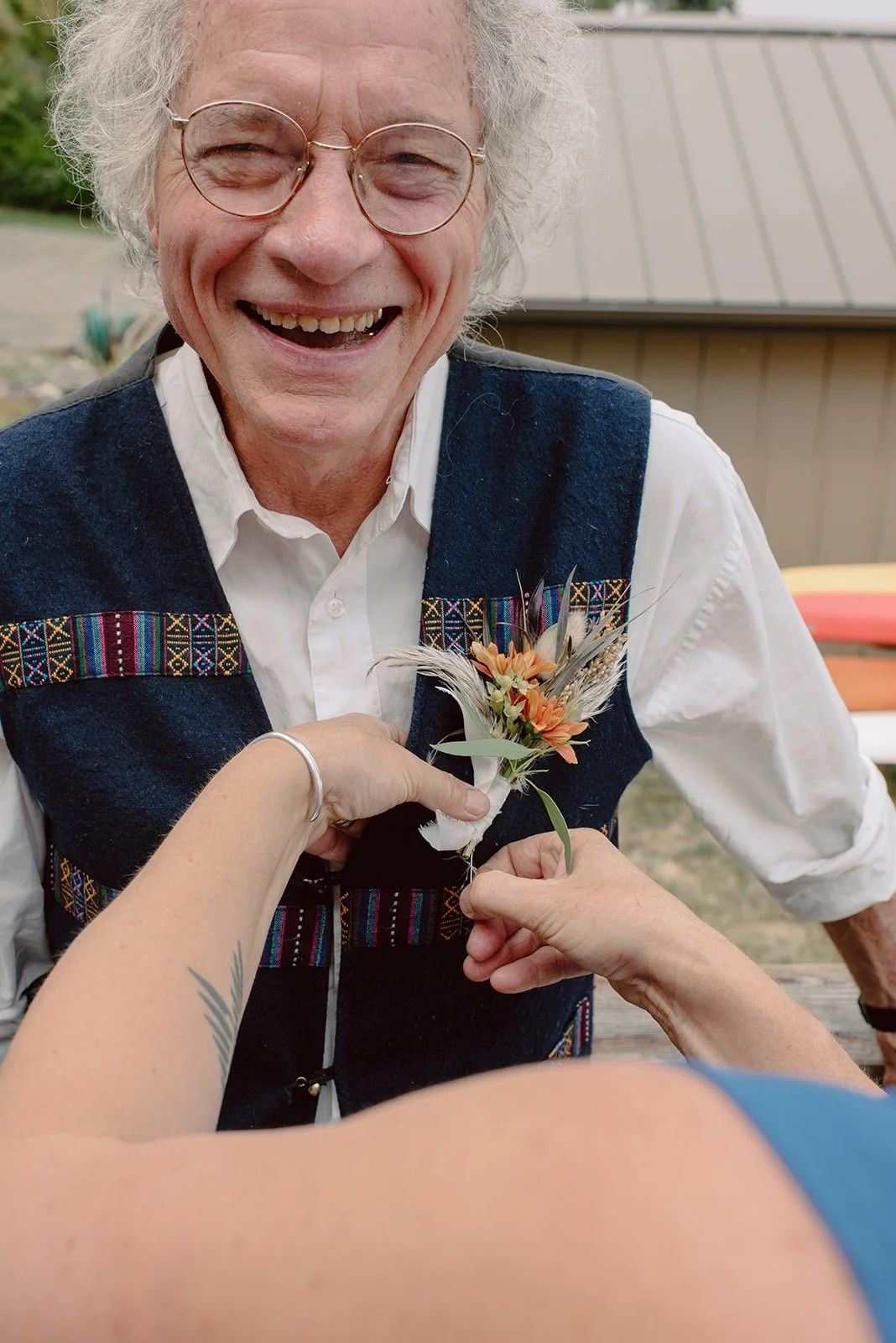 Smiling older gentleman getting a boutonniere put on his woven, blue wool vest. The boutonniere is made of orange chrysanthemums, purple grass, and dried elements.