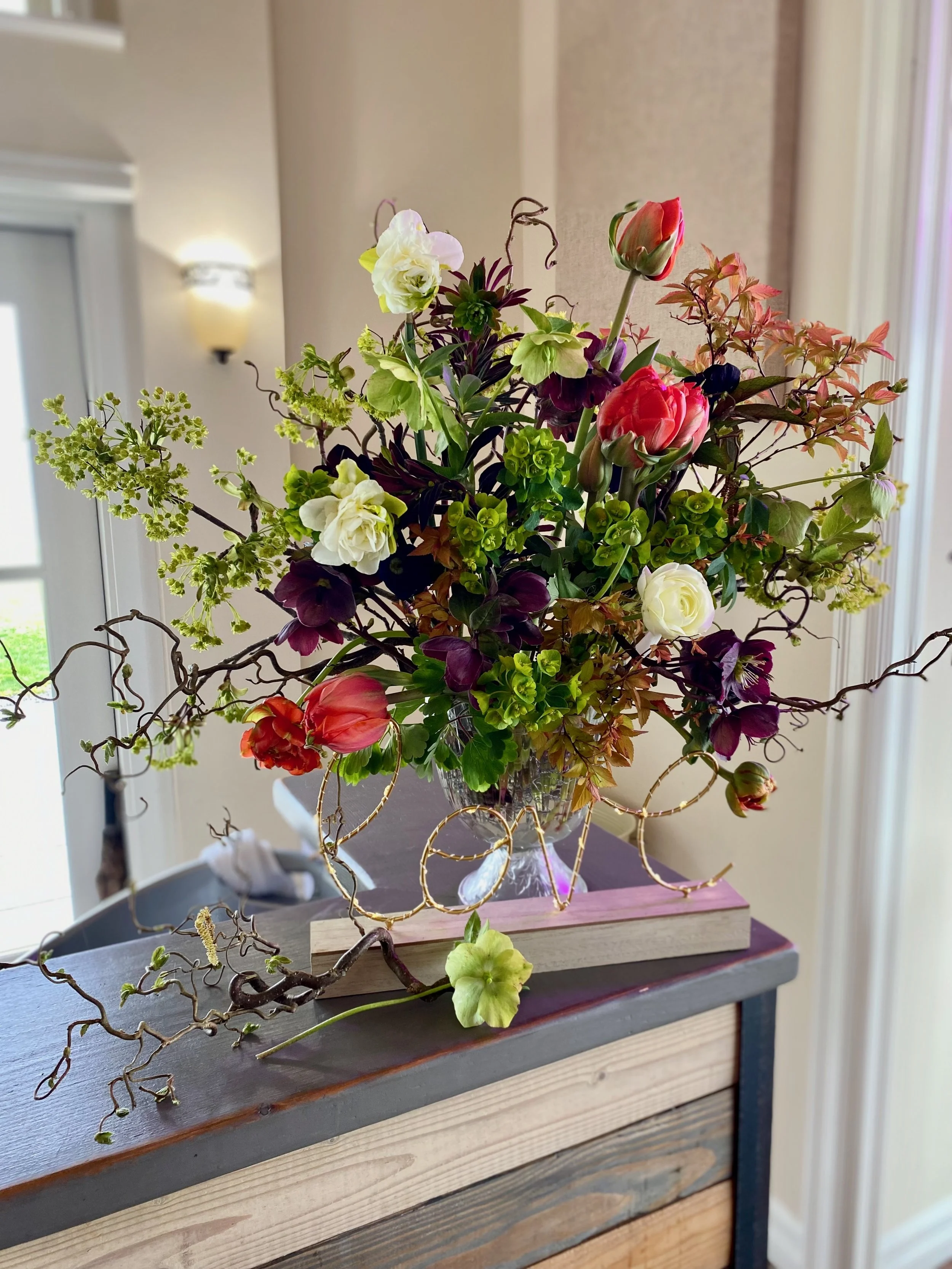 Large flower arrangement made with green and purple euphorbia, orange tulips, and fluffy white daffodils. Branches of curly hazelnut and Norway Maple in bloom add a fun textural element