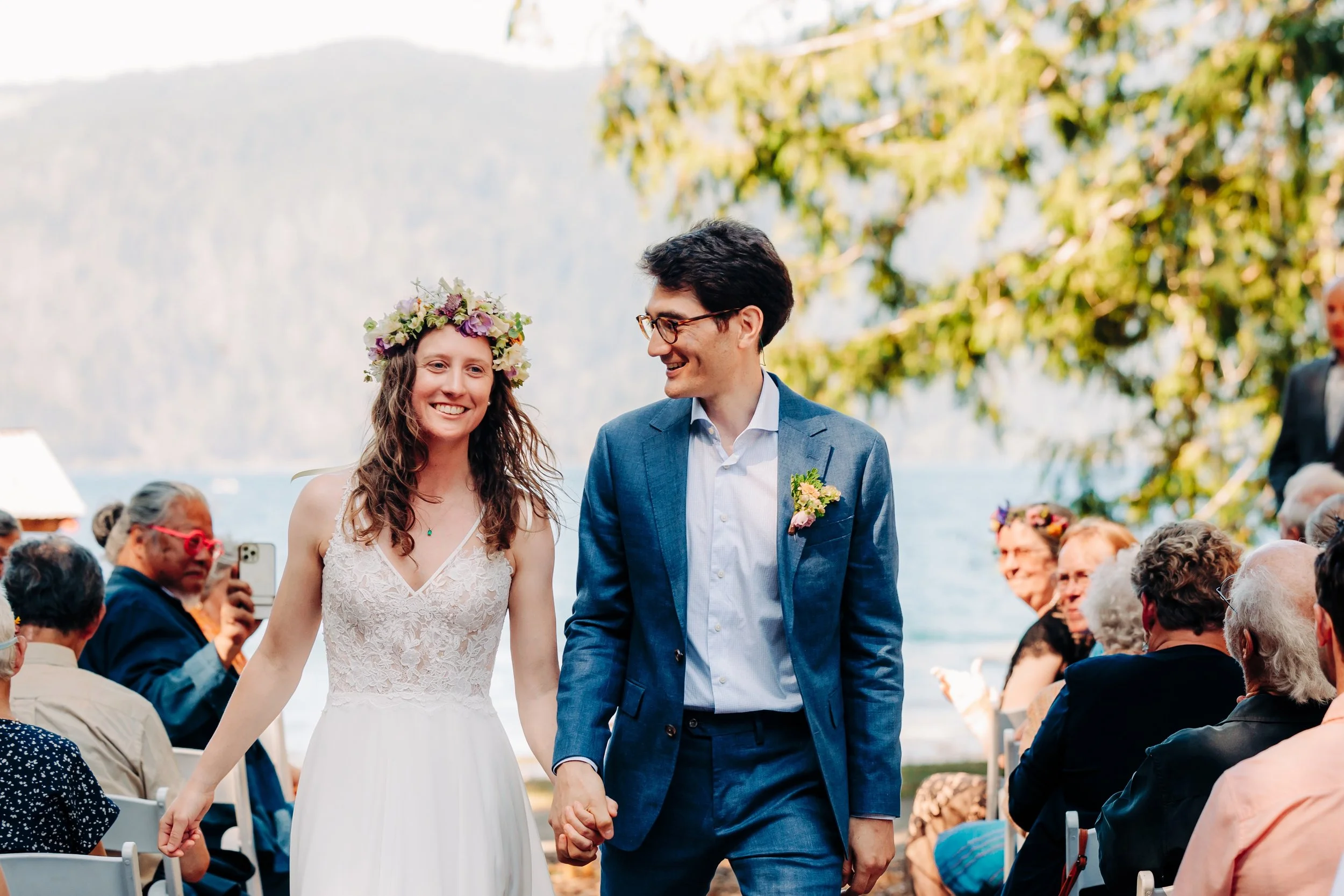 Couple holding hands walking down the aisle smiling after getting married. Outdoor wedding at NatureBridge on Lake Crescent. Spring colors flower crown.