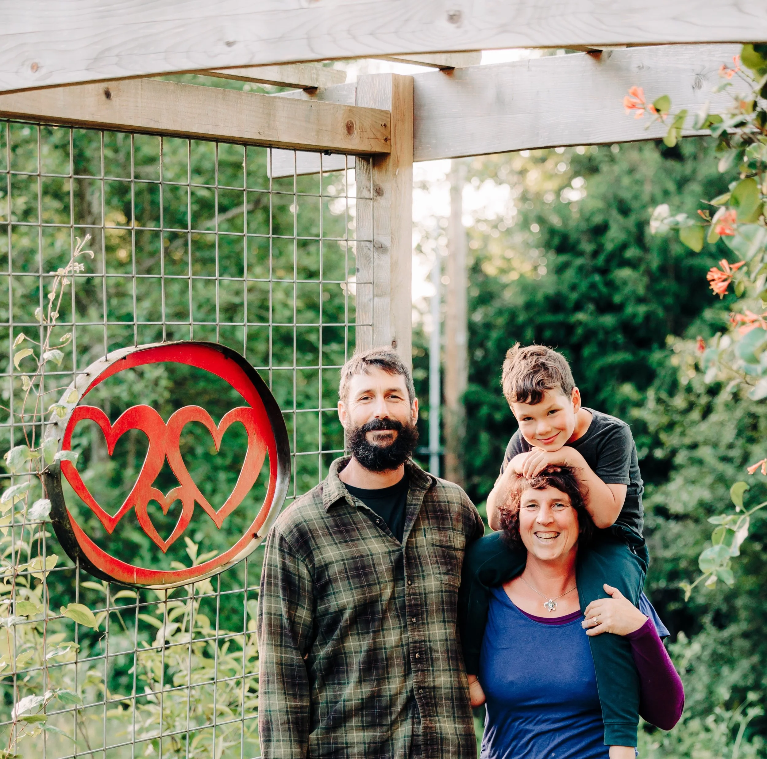 This is a photo of myself, my husband, and our young (6 years old) son. We are standing beneath a cedar arbor with a metal art piece with three red hearts (representing the three of us).