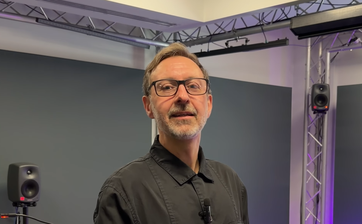 A man with glasses, a beard, and a mustache wearing a black shirt standing in a room with sound equipment and purple lighting.