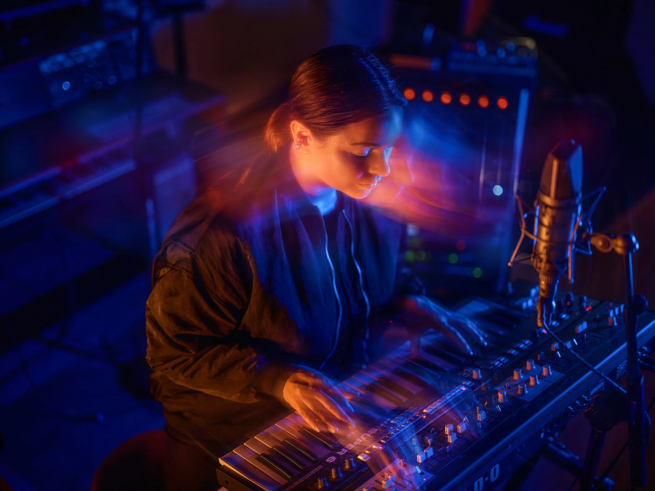 A female musician with dark hair in a ponytail playing a keyboard in a dark studio illuminated by multicolored lights, with a microphone set up nearby.